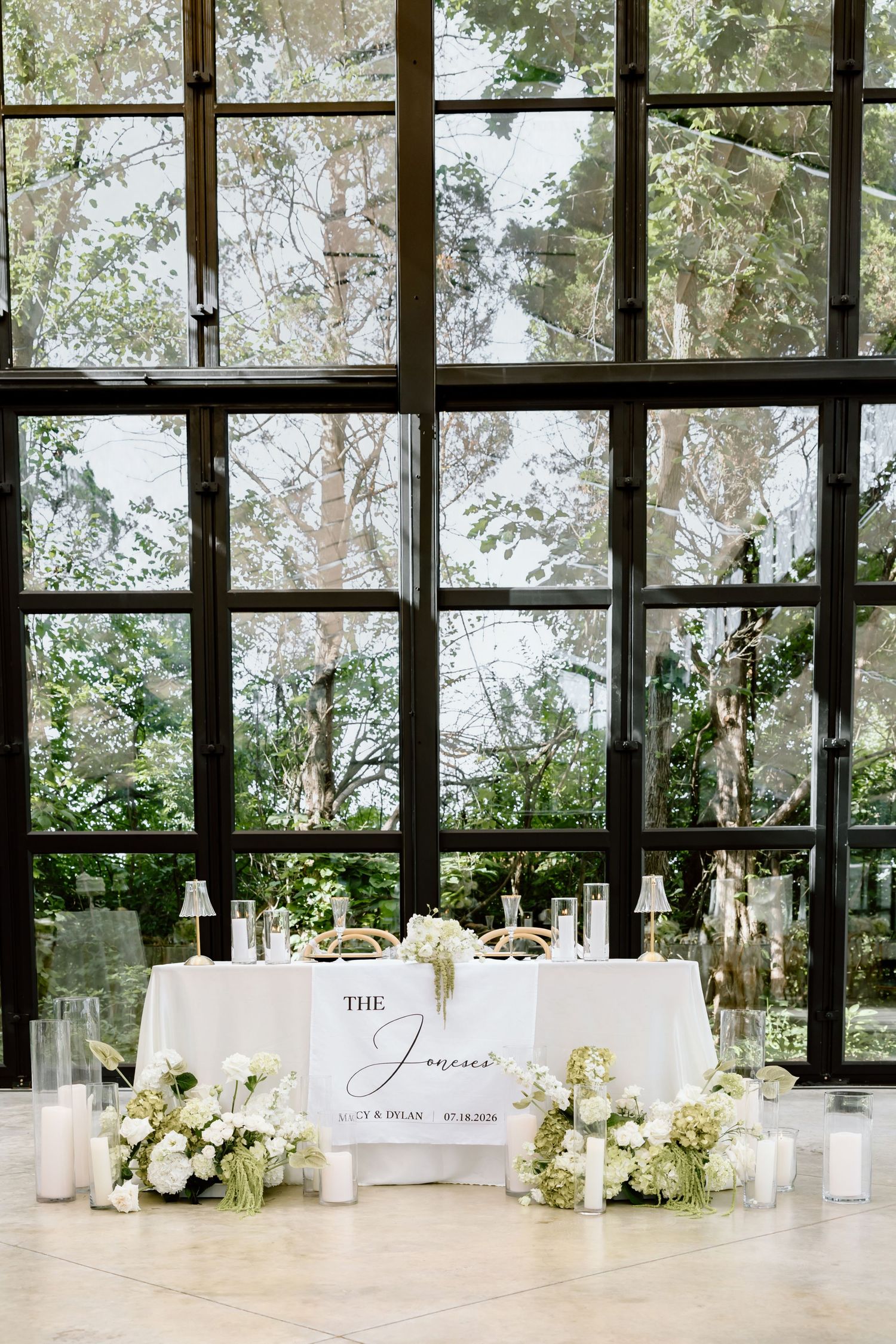 Elegant wedding sweetheart table with white florals, candles, and personalized sign against floor-to-ceiling windows.