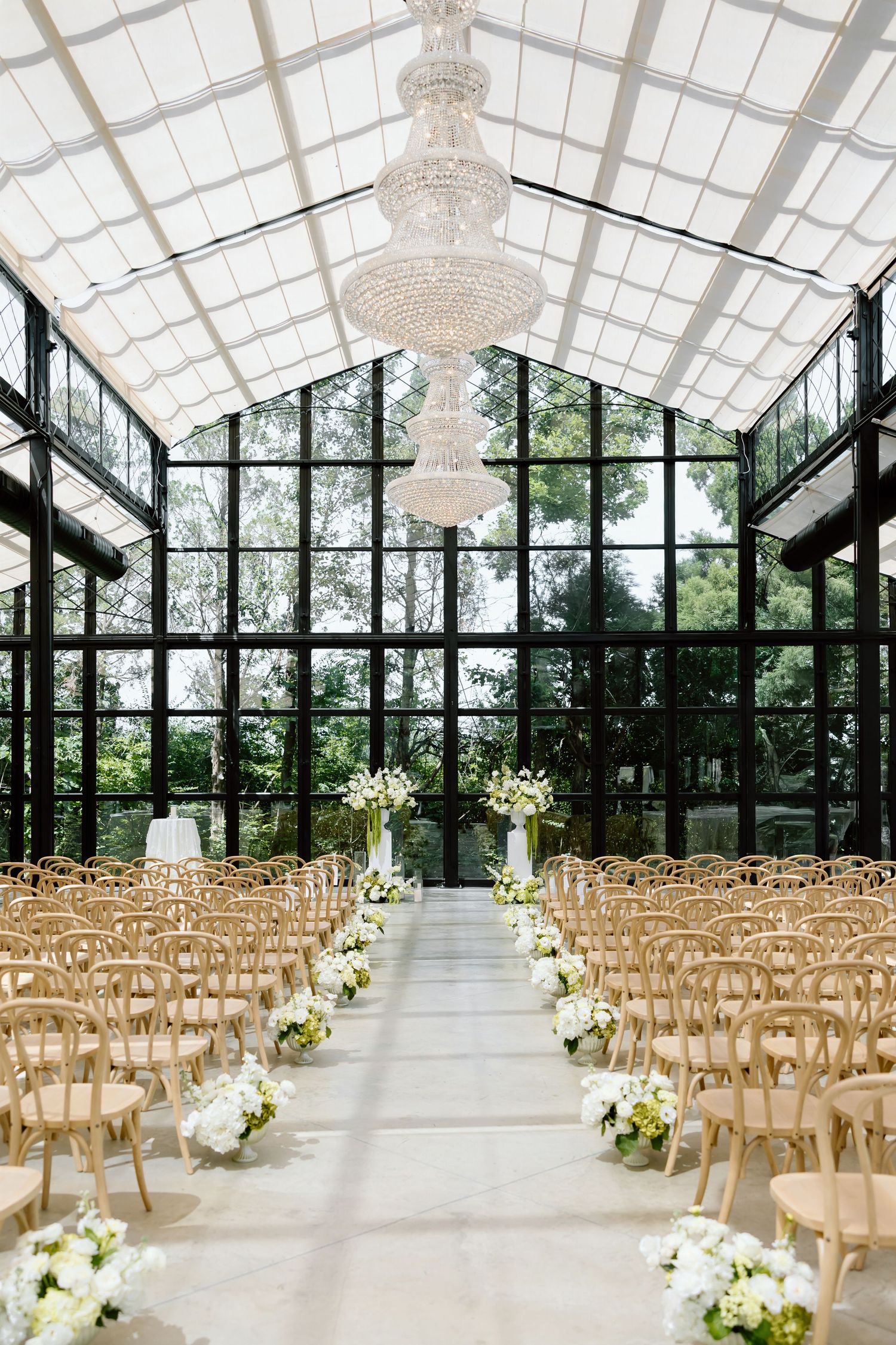 Elegant greenhouse wedding venue with crystal chandelier, wood chairs, white floral aisle decor, and floor-to-ceiling glass walls.