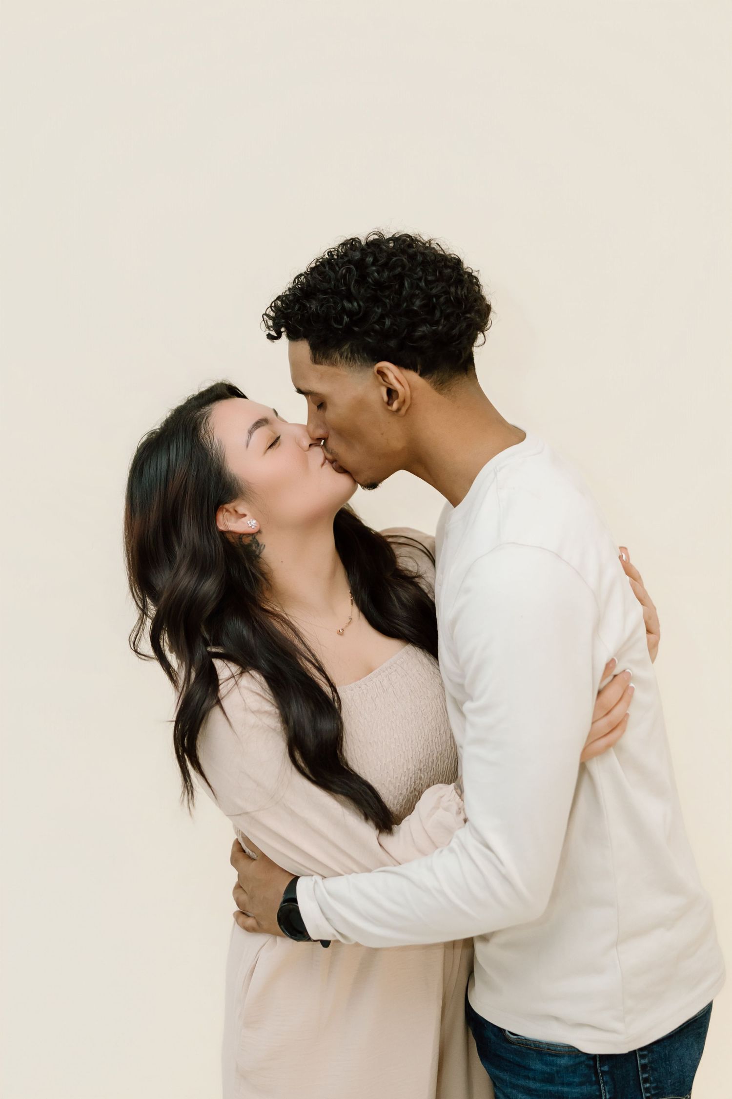 A romantic couple in neutral colored sweaters share a tender kiss against a light beige wall.