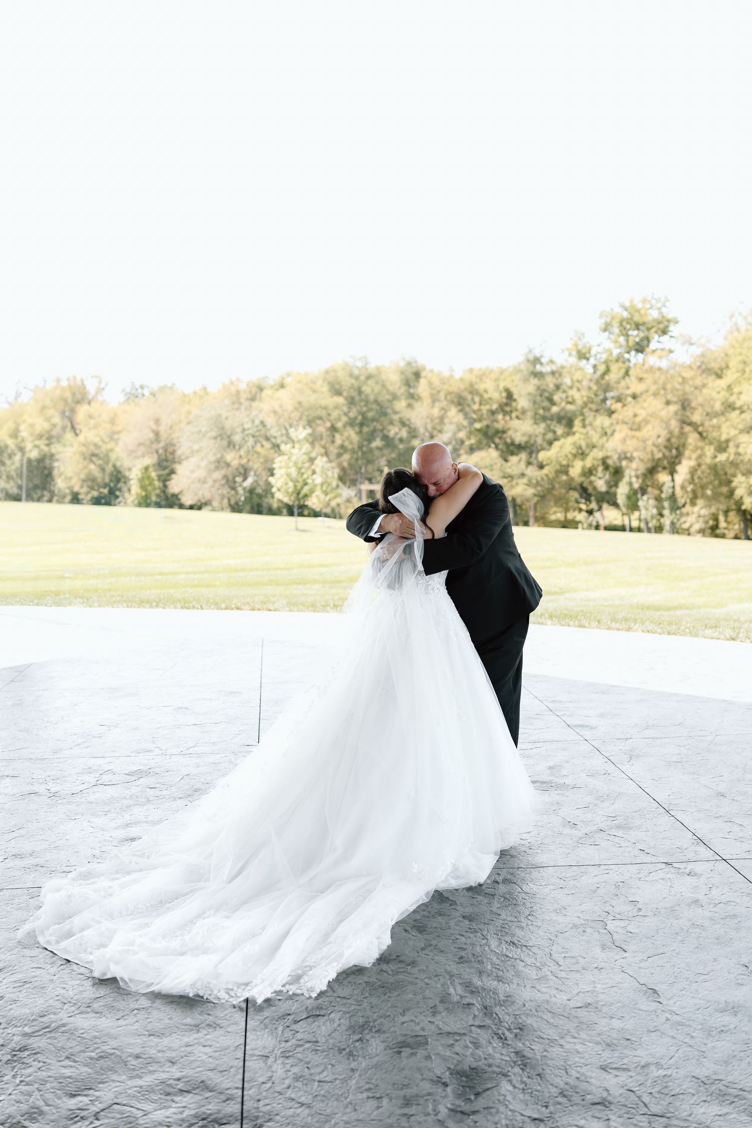 A couple shares a romantic kiss on their wedding day on a concrete patio with trees in the background.