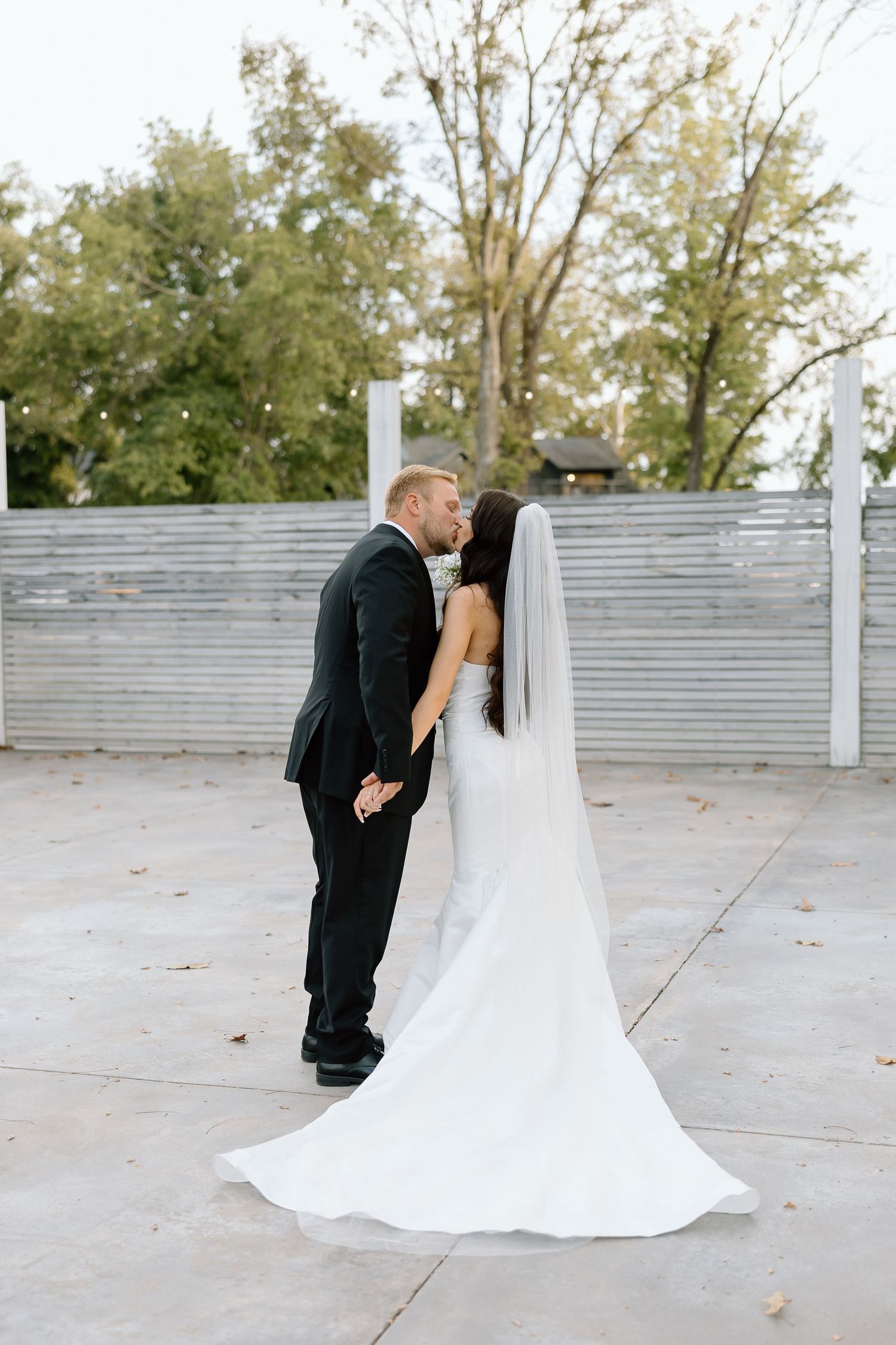 A romantic first look moment between newlyweds on an outdoor patio with white railings and fall trees in the background.