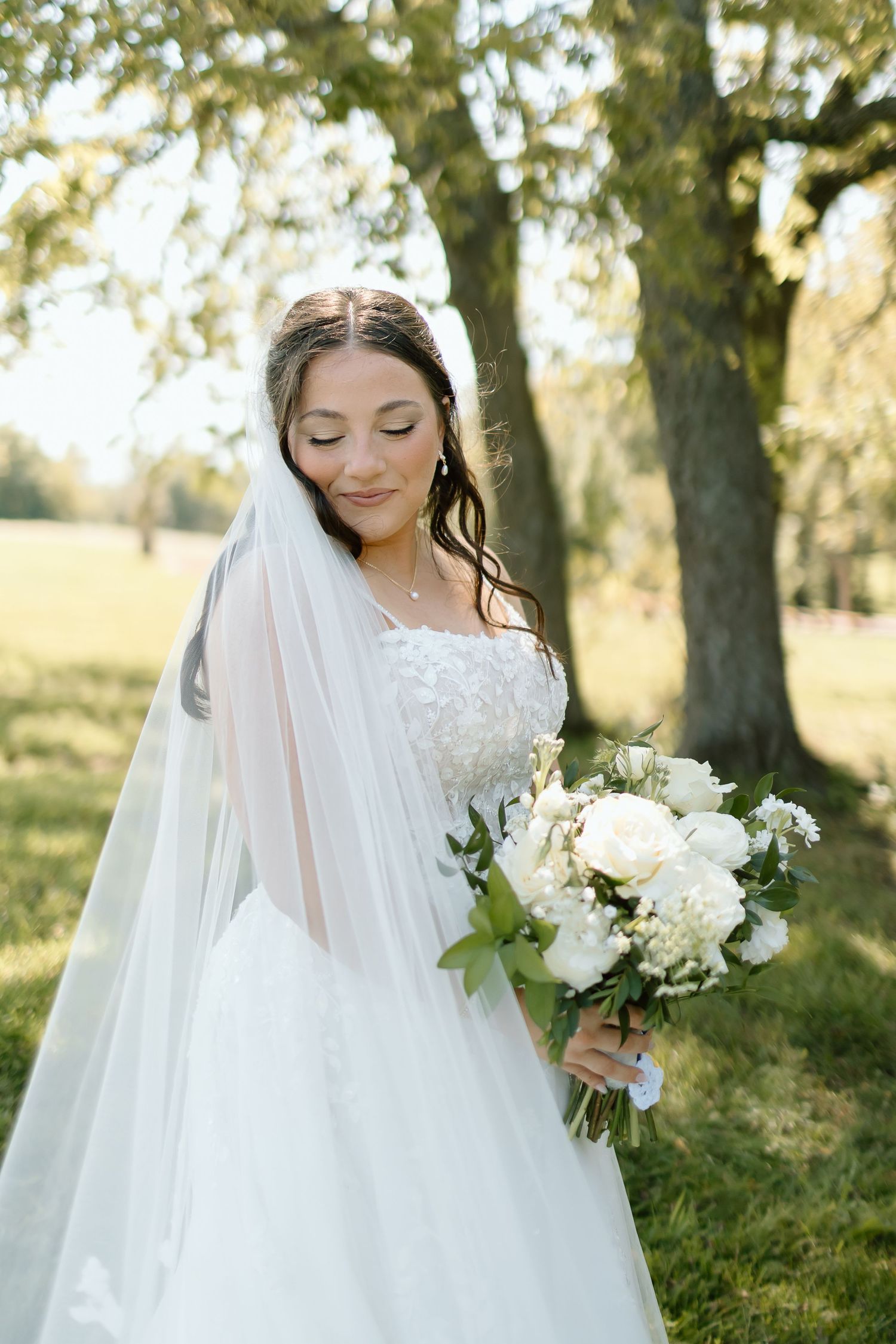 A bride in a white lace wedding gown with flowing veil holds an elegant white floral bouquet in a sunlit garden setting.