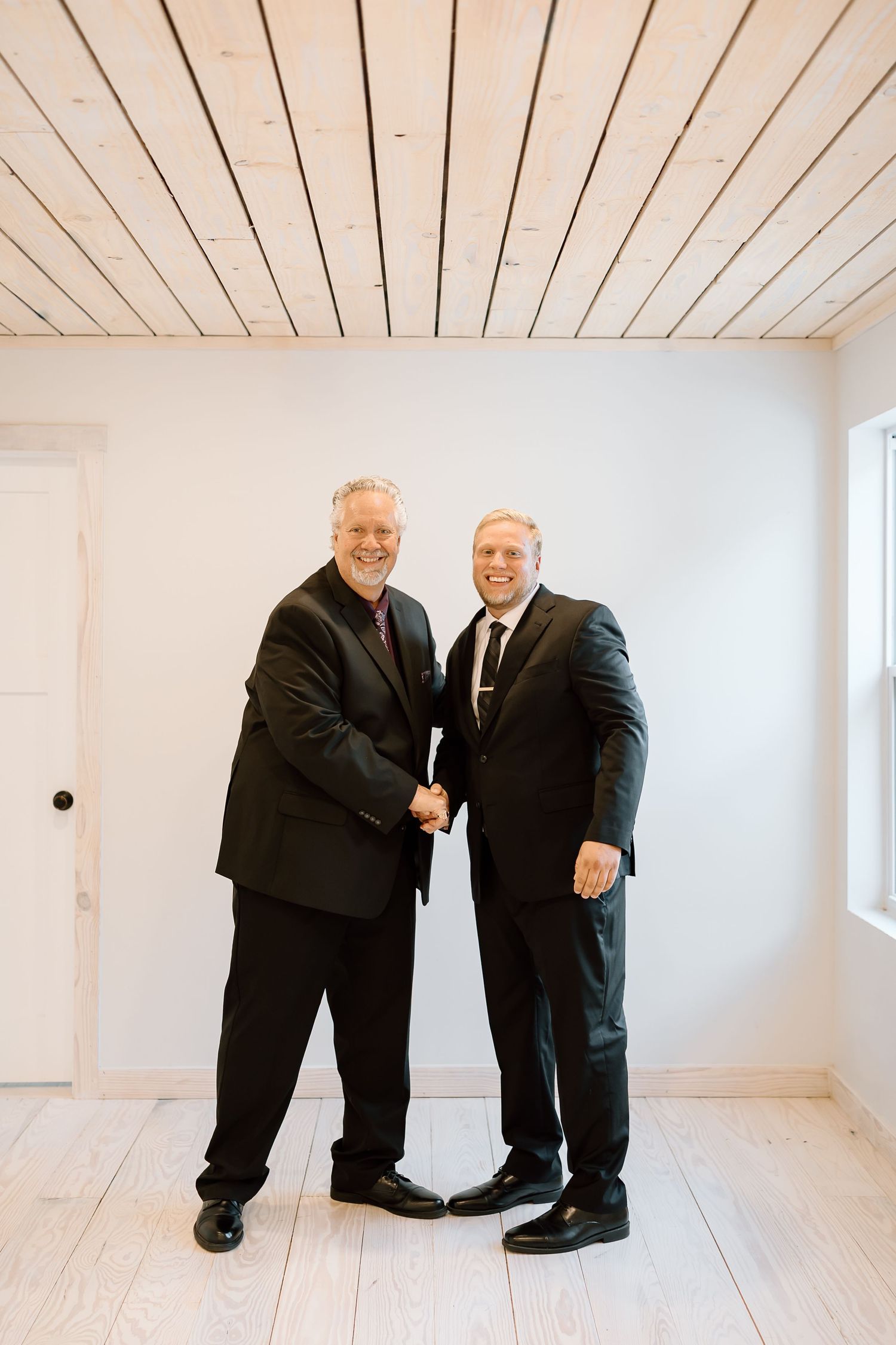 Two people in dark suits shake hands in a bright white room with wooden floors and ceiling.