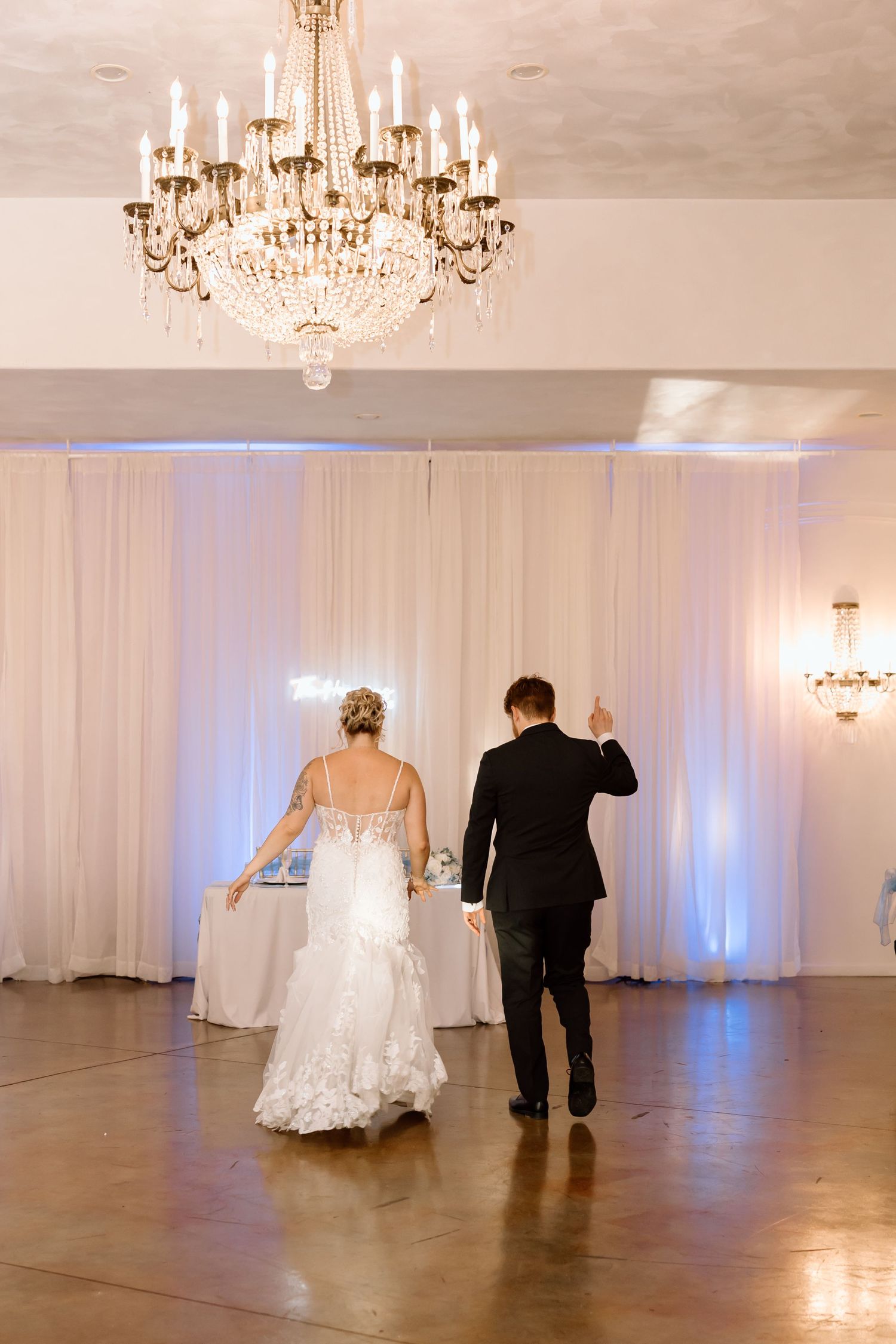 A couple in formal wedding attire walks across an elegant ballroom with crystal chandeliers and white drapery.