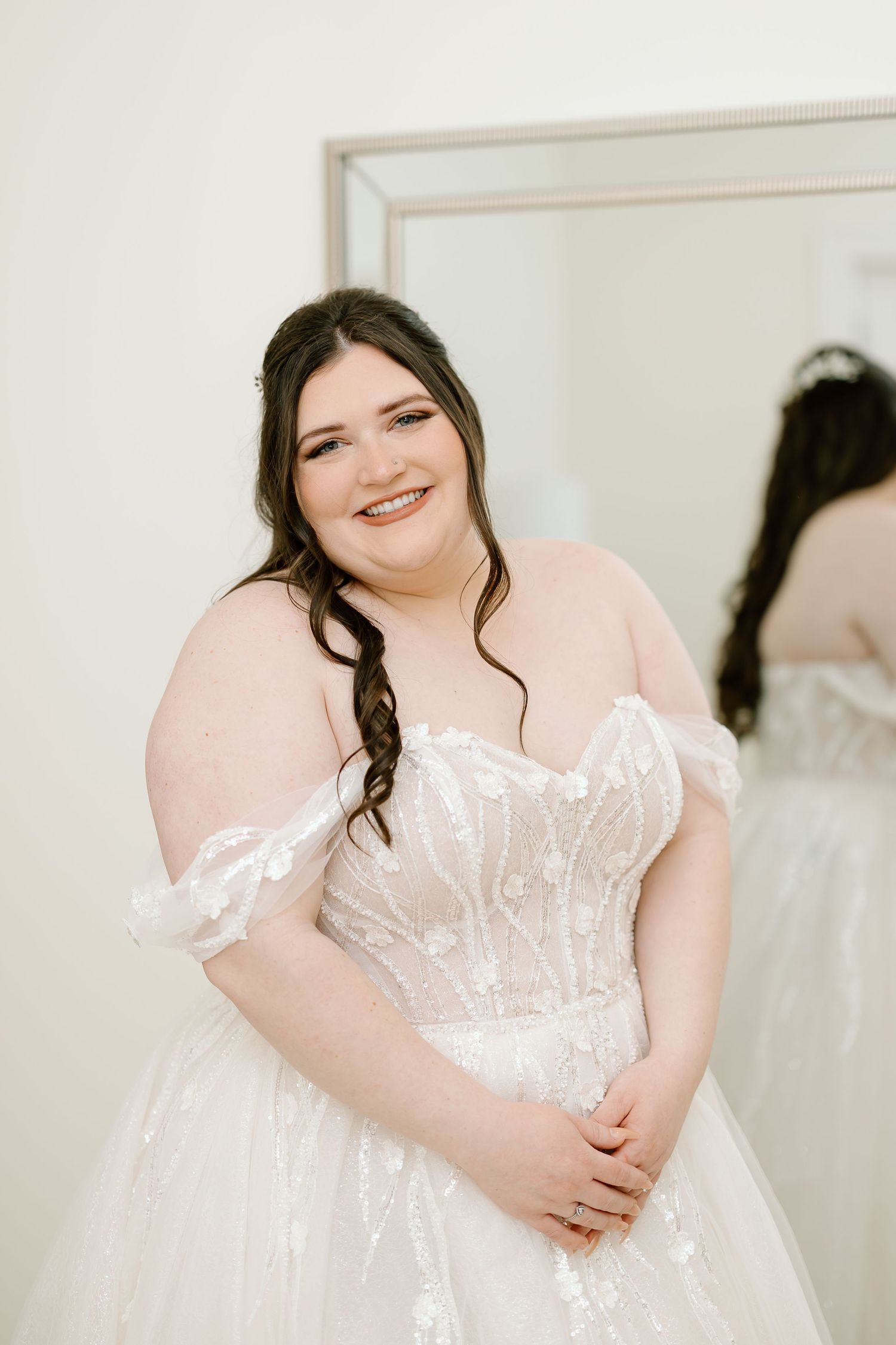 A plus size model poses gracefully in a white lace wedding gown with delicate off-shoulder details in a bridal boutique.