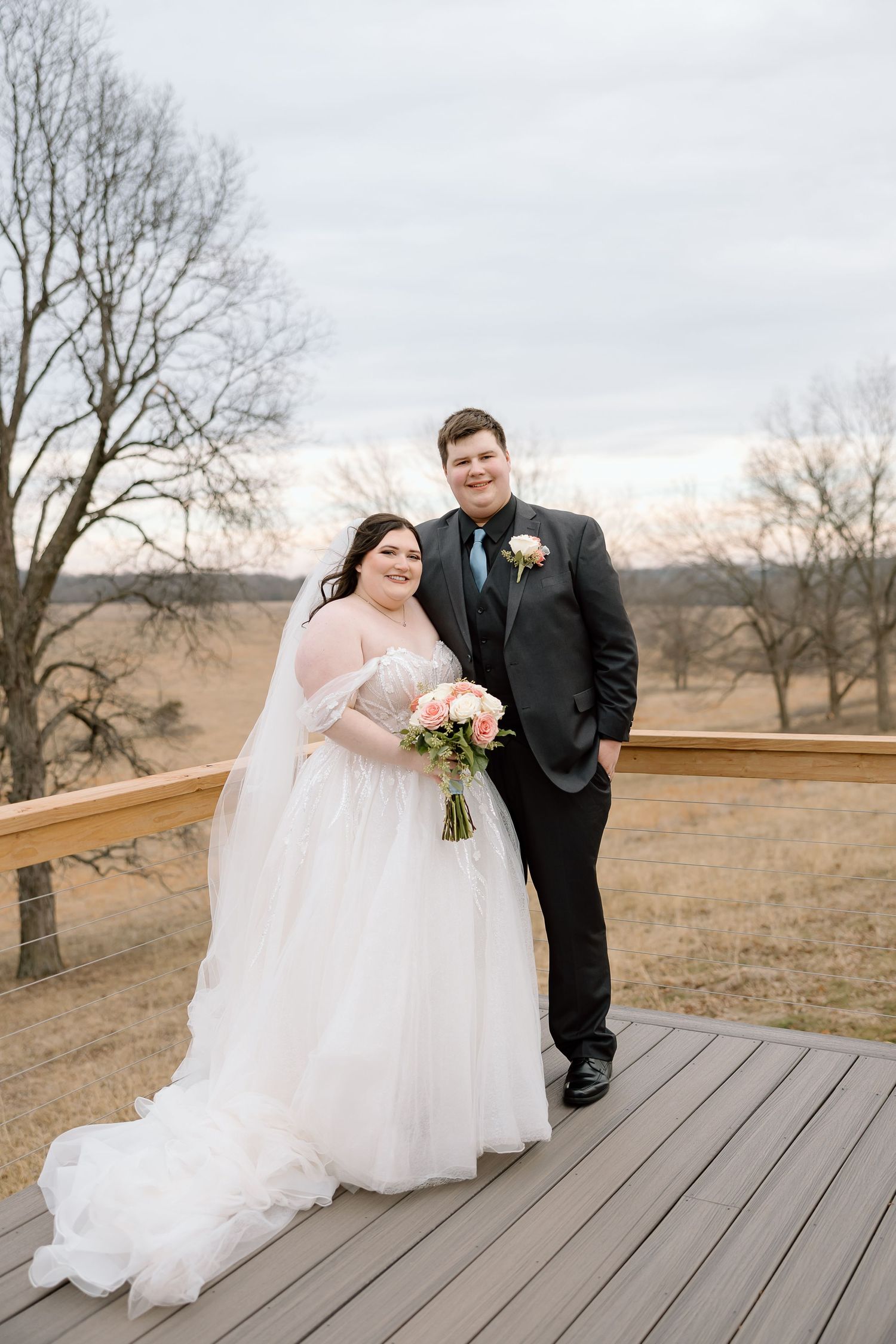 A couple in wedding attire sits together on a wooden deck overlooking a rustic winter landscape at sunset.