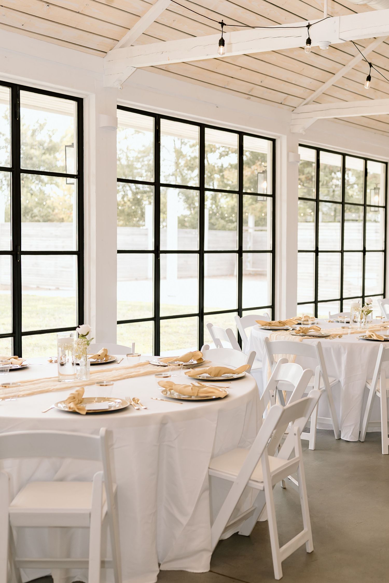 A bright white wedding reception venue with large windows, wooden beams, and tables set with white linens and gold chargers.