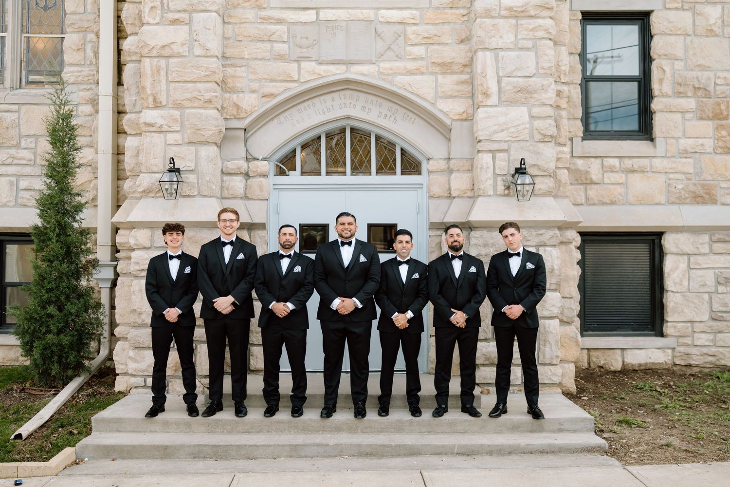 Seven groomsmen in black tuxedos and bow ties pose on steps outside a stone church wedding venue.