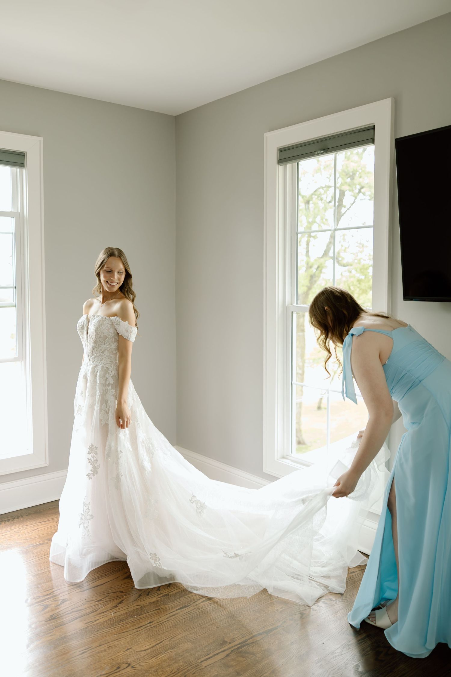 Bride in white lace gown with bridesmaid in blue dress arranging the train in a bright room.
