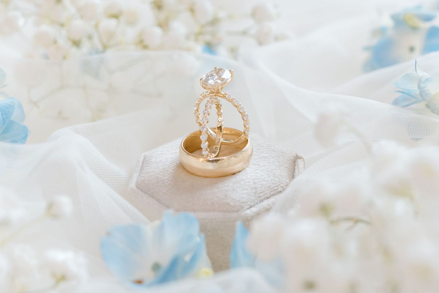 Gold wedding bands and diamond engagement ring displayed on a small pillow surrounded by white and blue flowers.