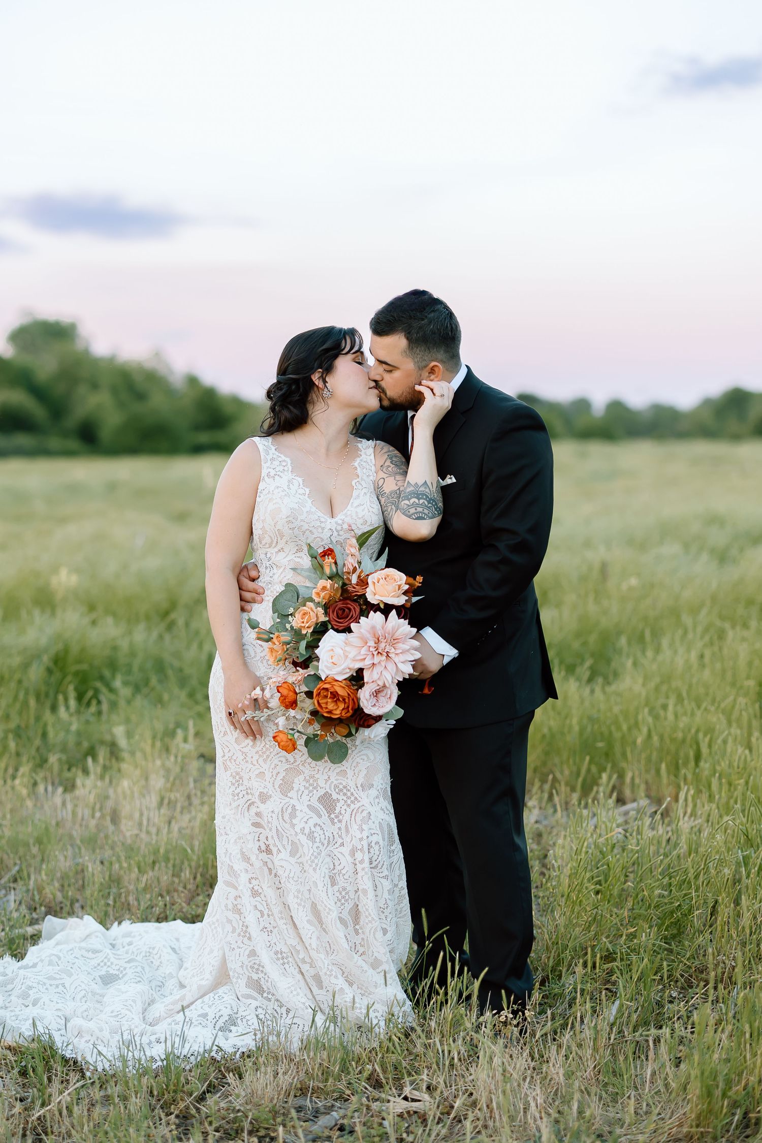 A romantic couple in wedding attire shares tender moments together in a sun-lit grassy meadow field.