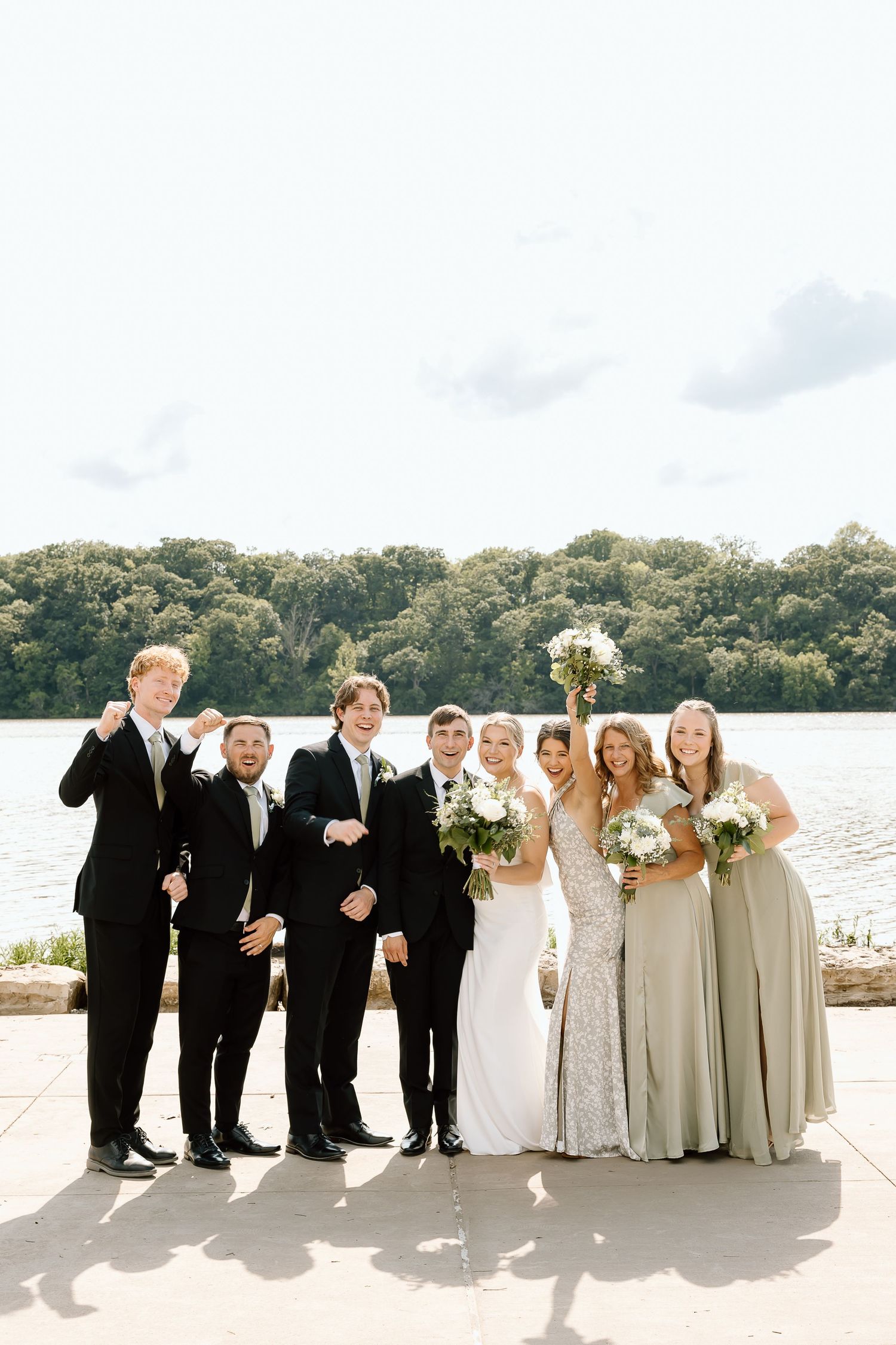 Wedding party enjoys a candid moment by the waterfront, groomsmen in black suits and bridesmaids in light gowns.