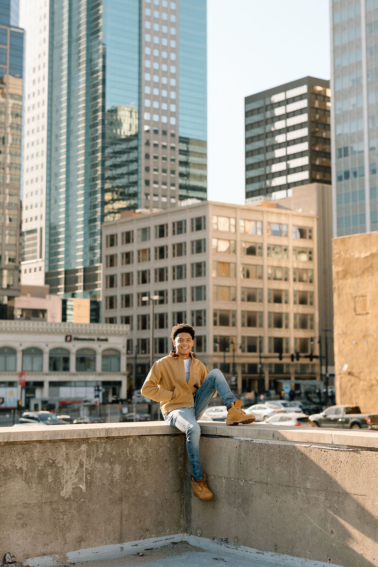 Person in tan jacket sits casually on concrete ledge with city skyline backdrop.