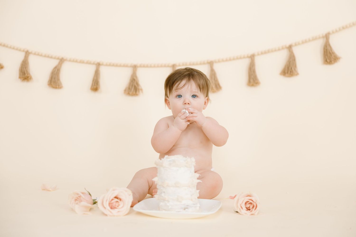 A baby covers mouth while sitting with white cake and pink roses under tassel banner.