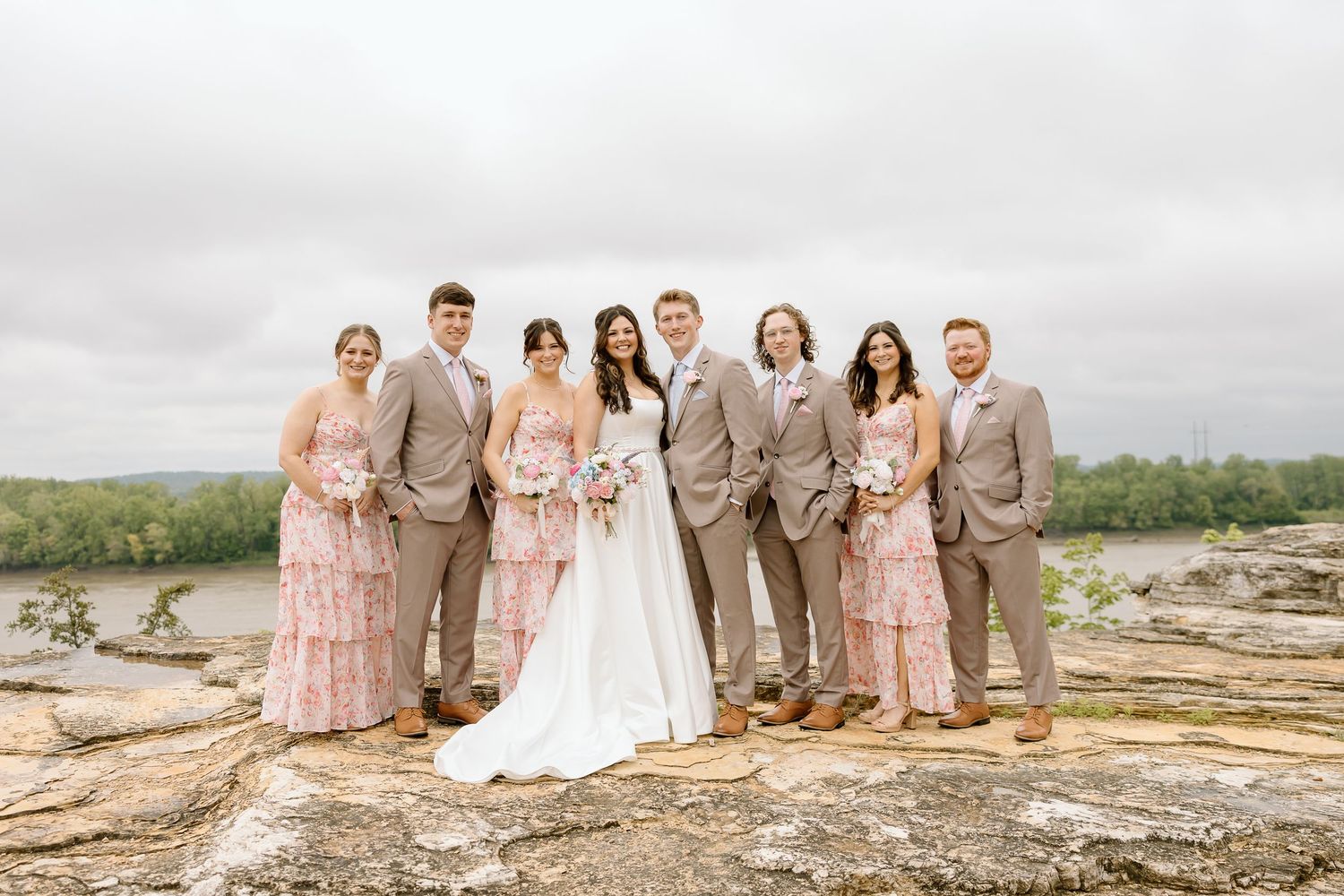 Wedding party in coordinated pink and tan attire poses on rocky overlook with water view.