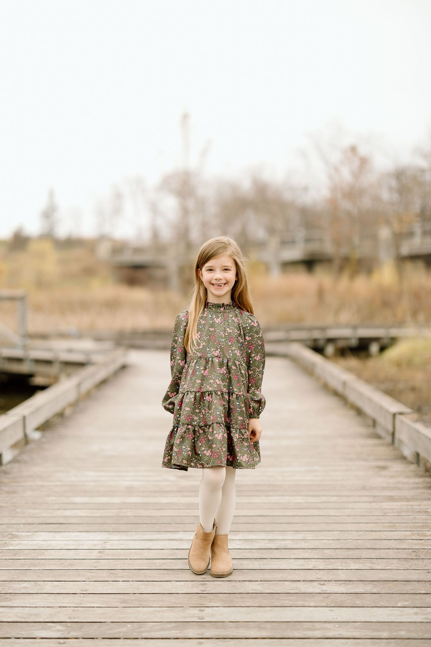 A child in a floral dress stands alone on a wooden boardwalk during golden hour.
