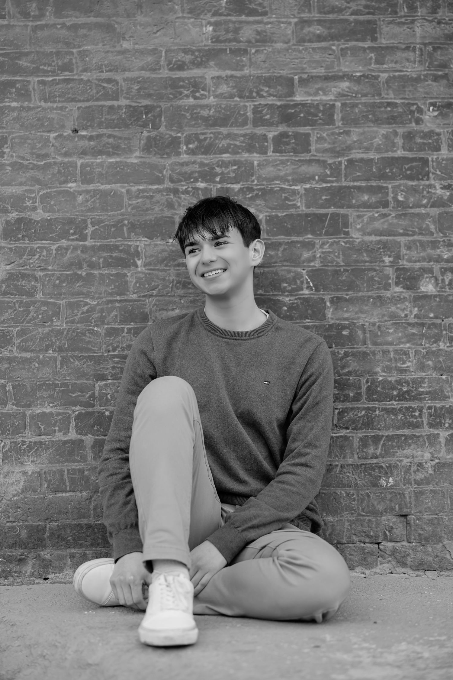 Black and white image of person in casual attire sitting against brick wall.