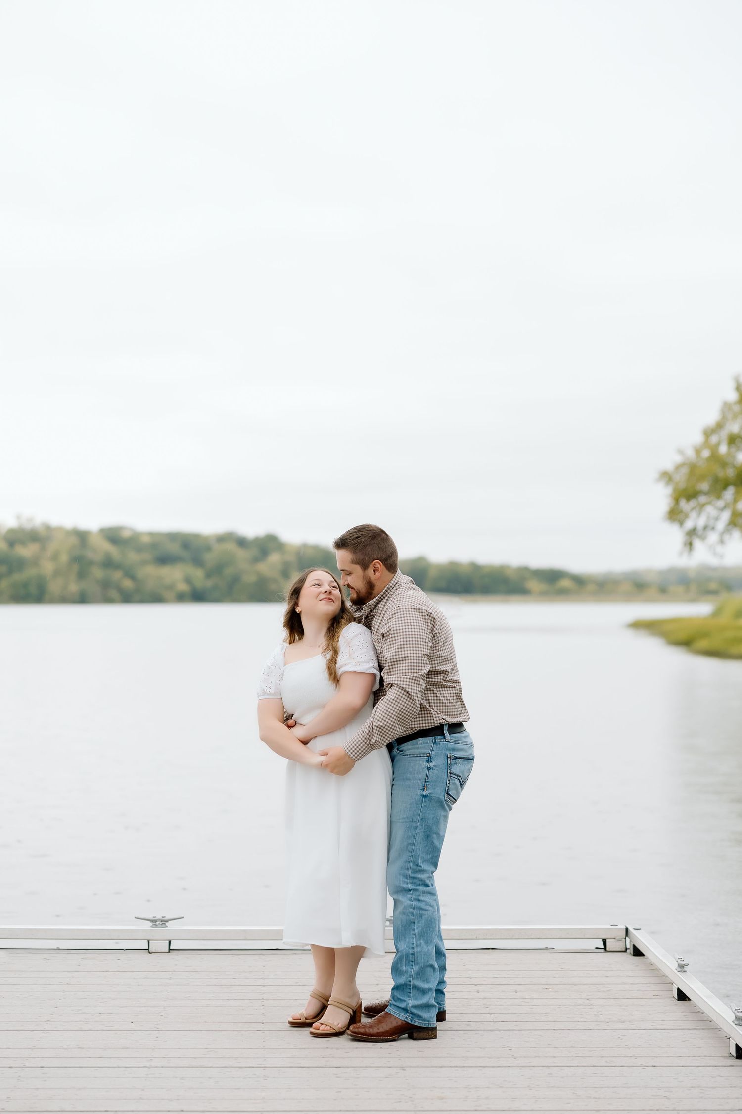 A couple embraces tenderly on a lakeside dock during an overcast day.