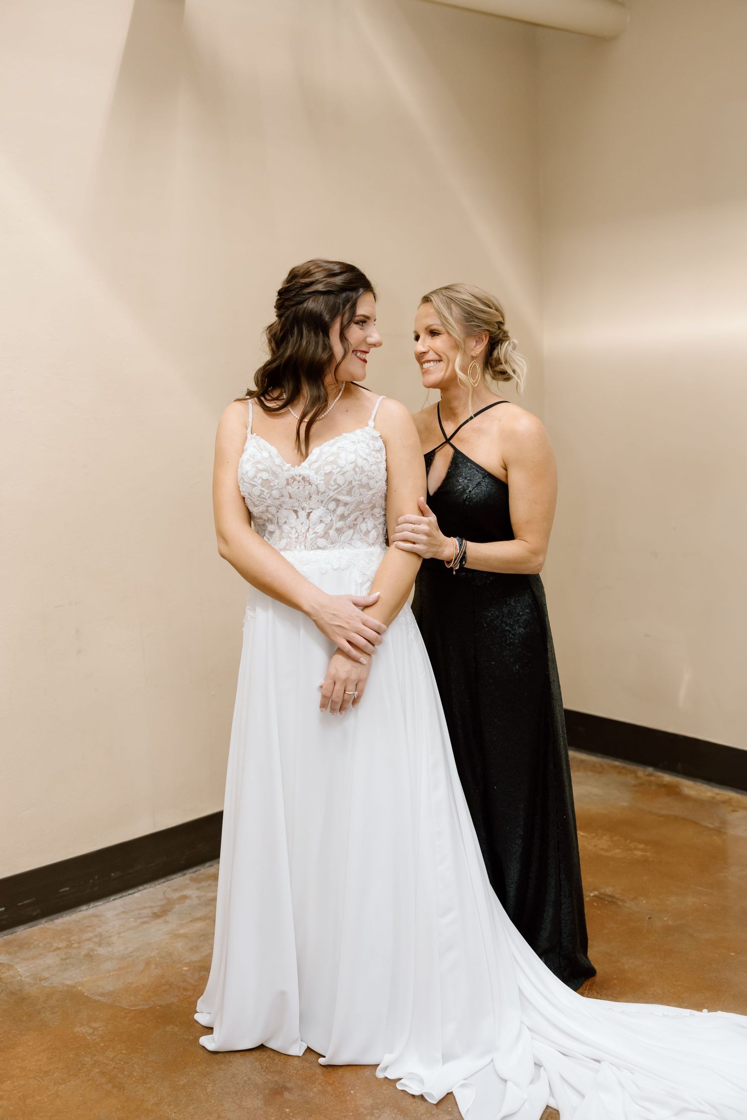 Two women in formal gowns, one wearing white and one in black, share a joyful moment together in a hallway.