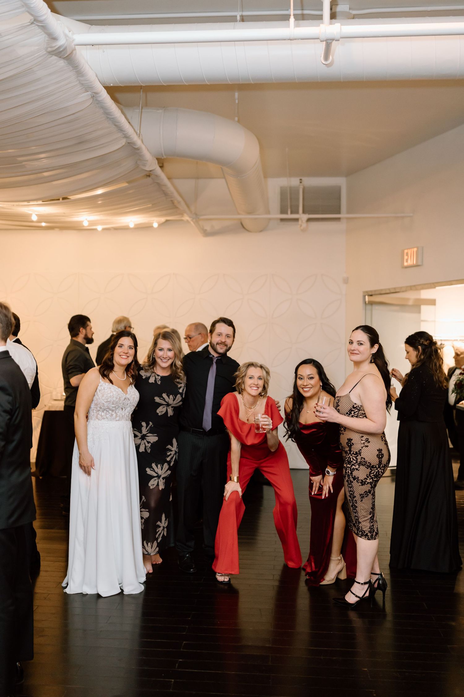 Wedding guests dance and socialize on a dark wooden floor in an indoor reception venue with white walls.