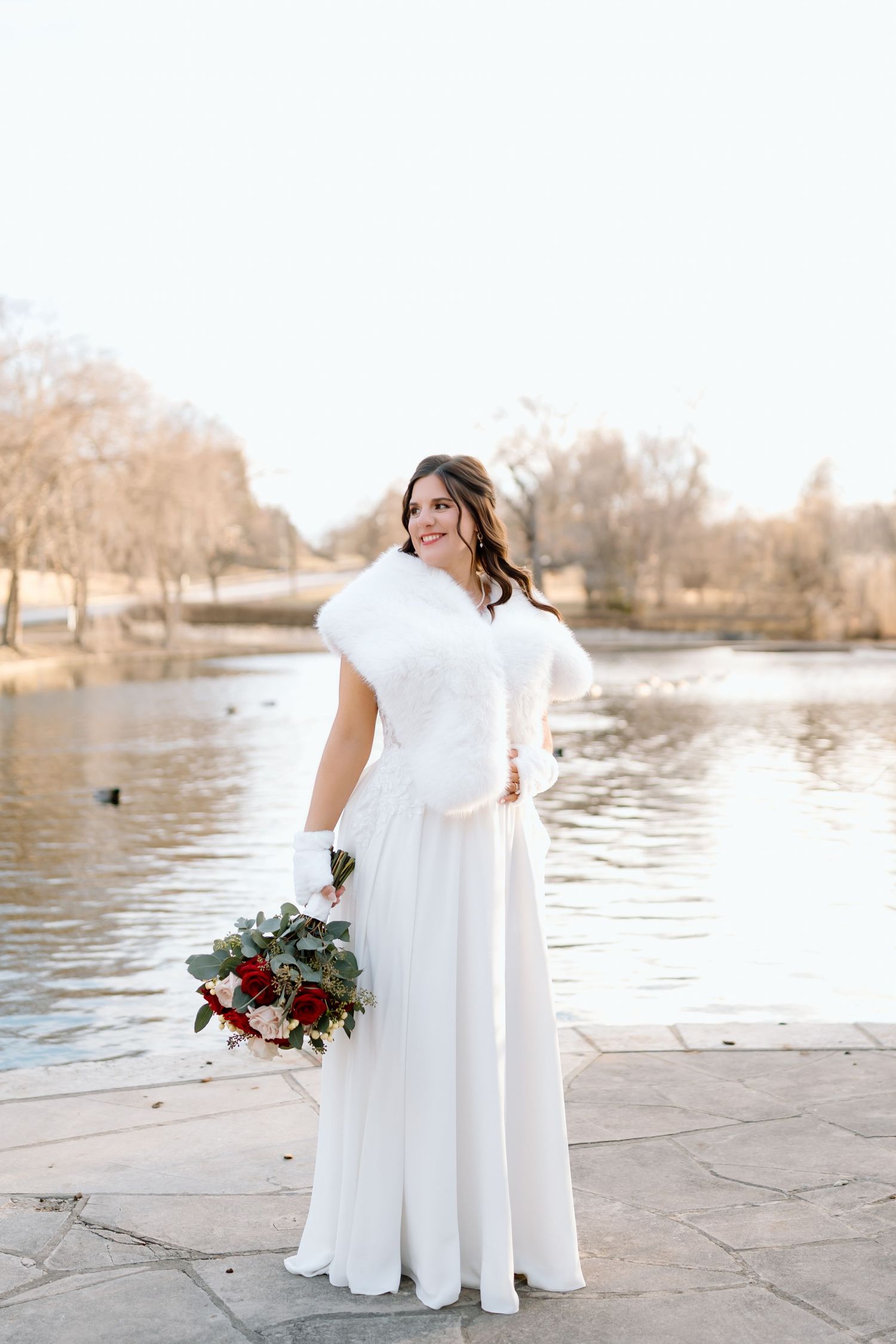 A bride in a flowing white dress stands by a serene lake during a winter photoshoot with burgundy florals.