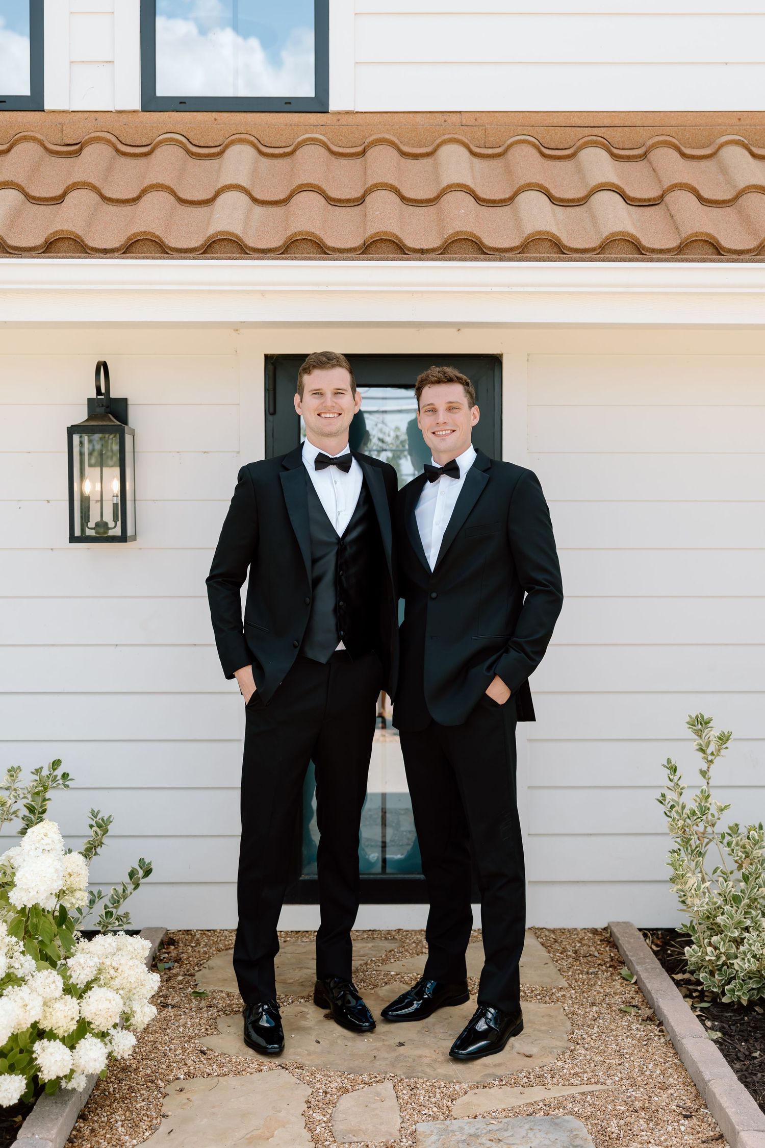 Two men in black tuxedos with bow ties smile outside a white building with hydrangeas.