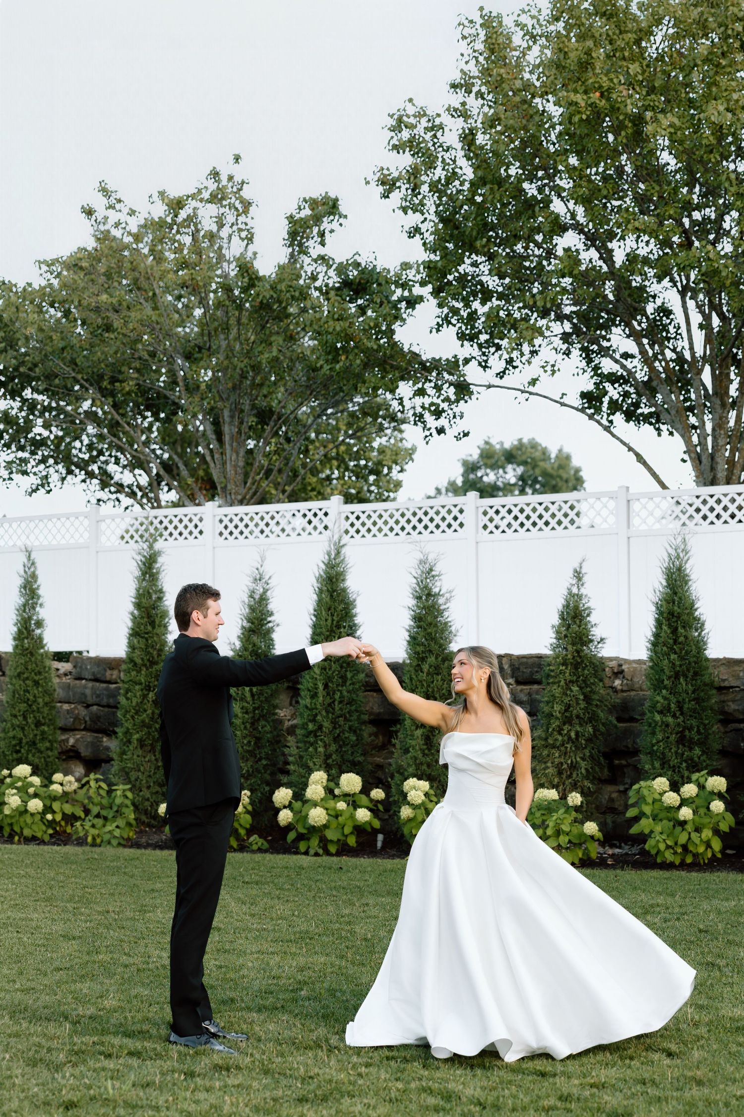 A bride in a white gown twirls as her groom in a black suit holds her hand in a lush garden setting.