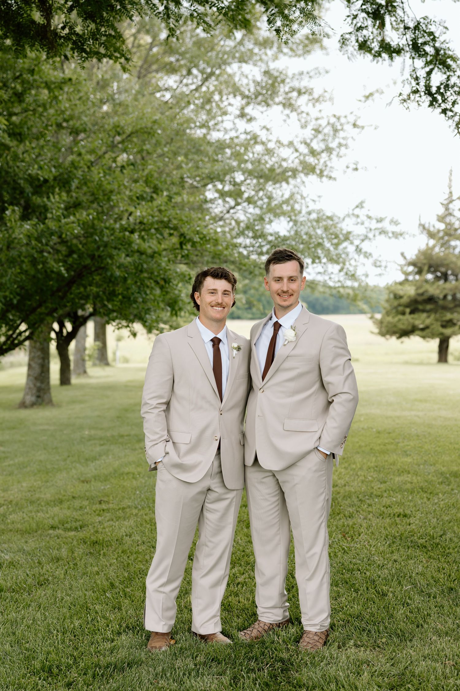 Two grooms in matching tan suits with brown ties smiling outdoors on green grass beneath shade trees.