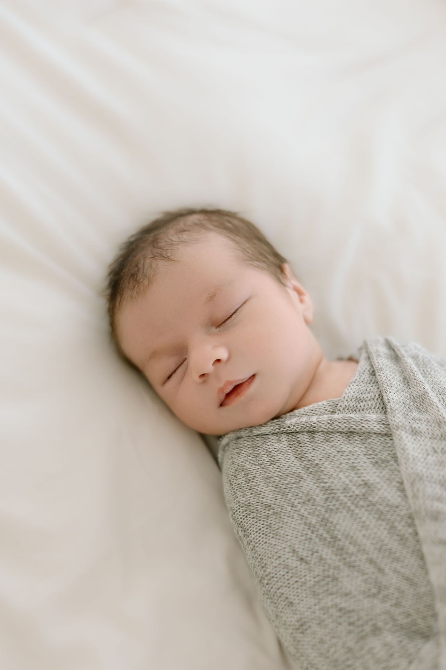 A sleeping newborn wrapped in a cozy gray knit blanket rests peacefully on white bedding.