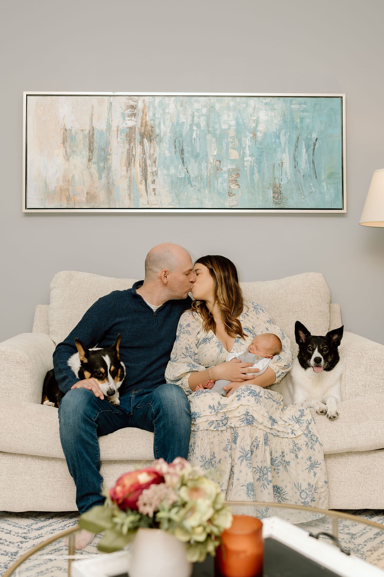 A couple sits on a white couch with their baby and pets while abstract art hangs on the wall behind them.