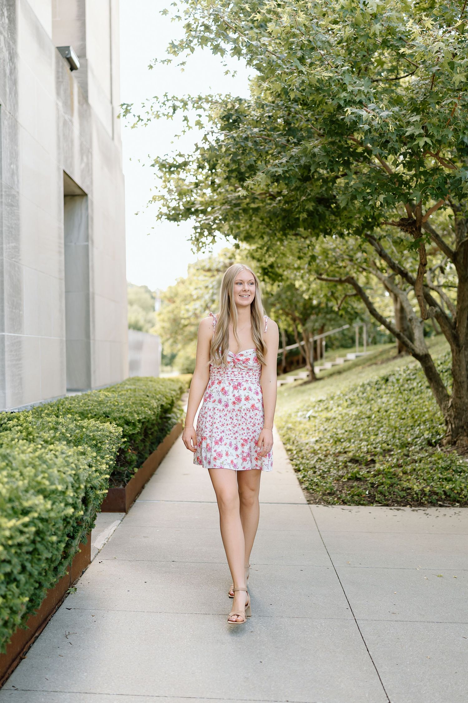 A person in a floral print dress walks along a concrete path surrounded by green landscaping and architectural columns.