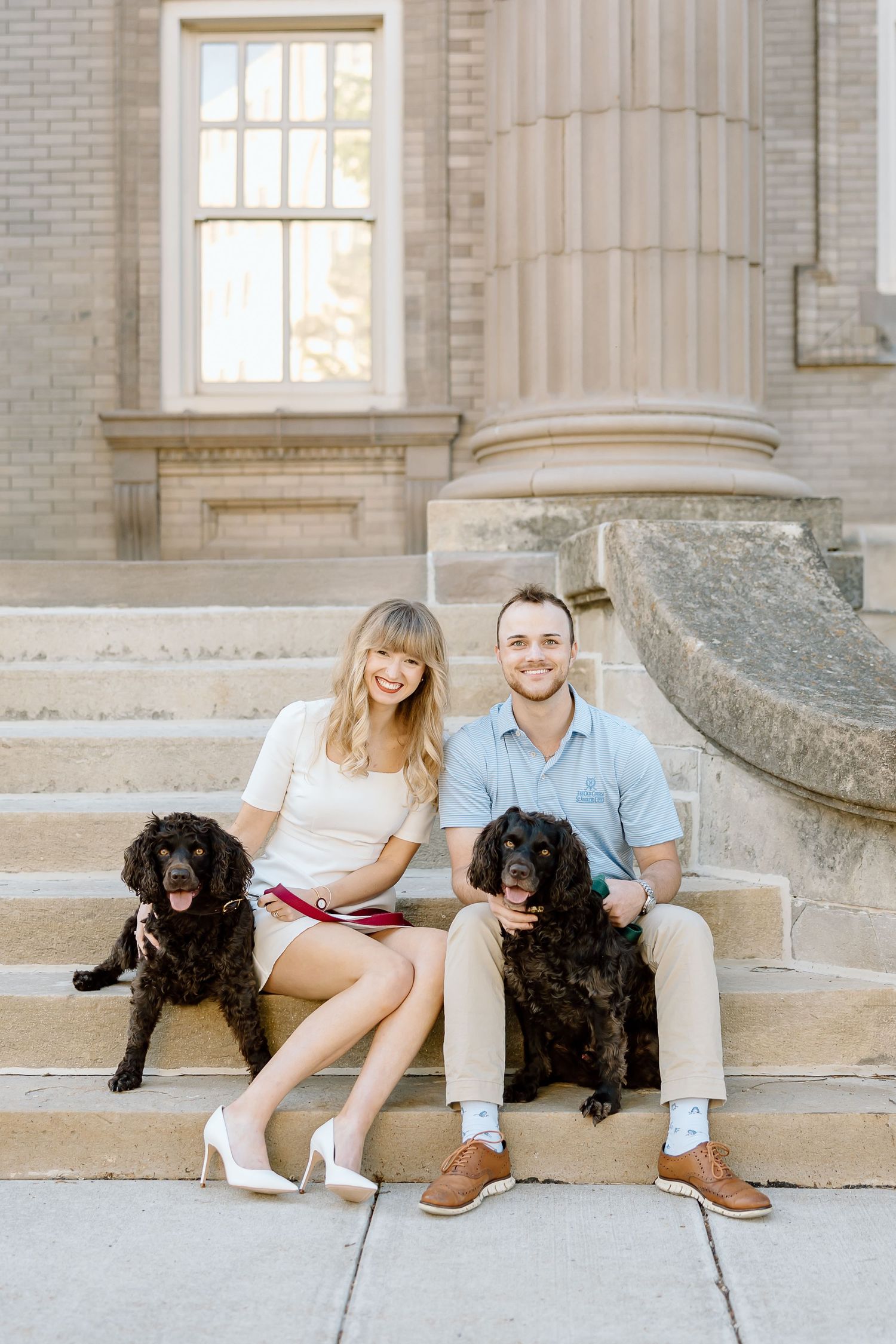 A couple sits on stone steps with their two black Newfoundland dogs outside a historic building with columns.