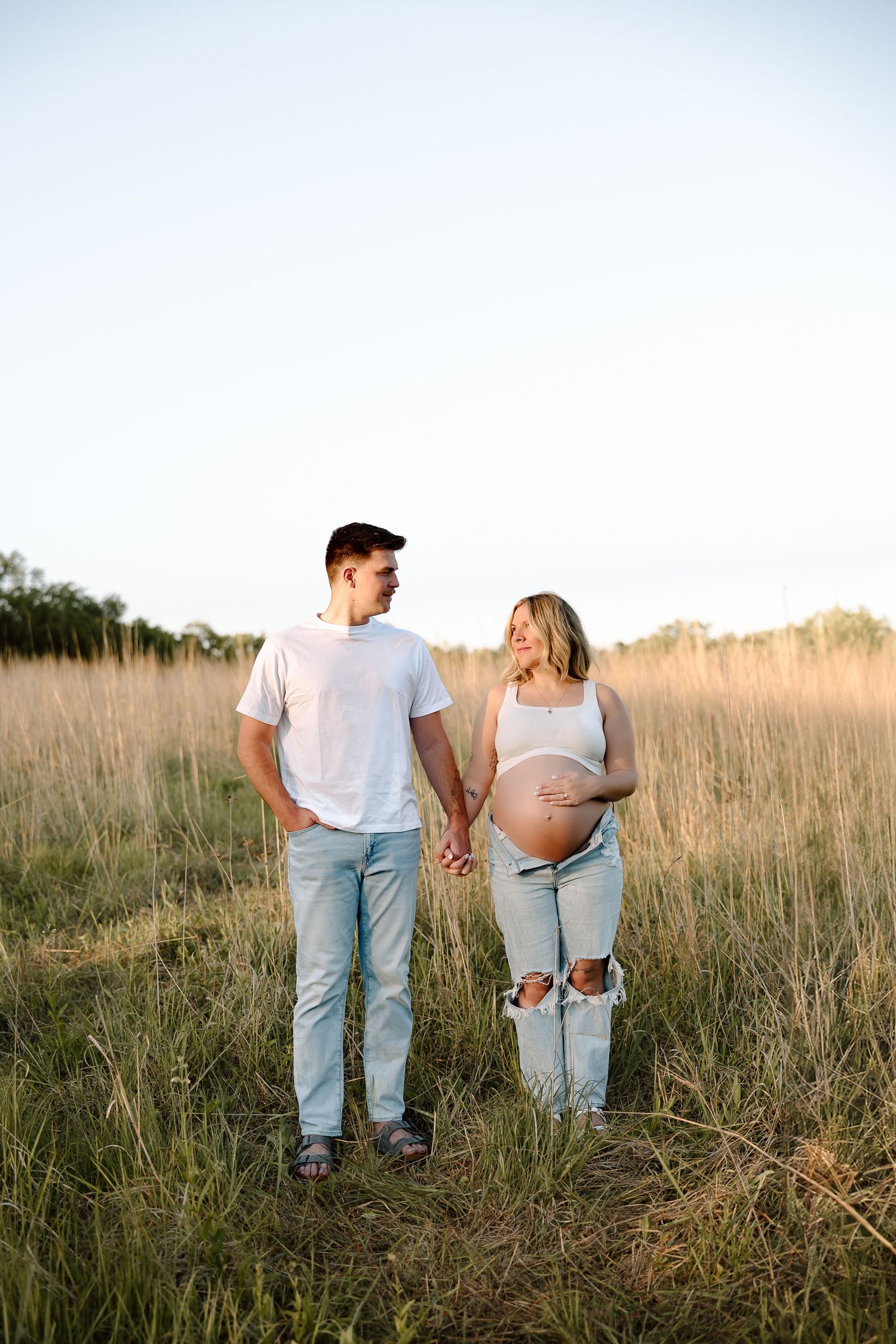 A couple stands together in a golden field during sunset, with the pregnant woman showing her baby bump.