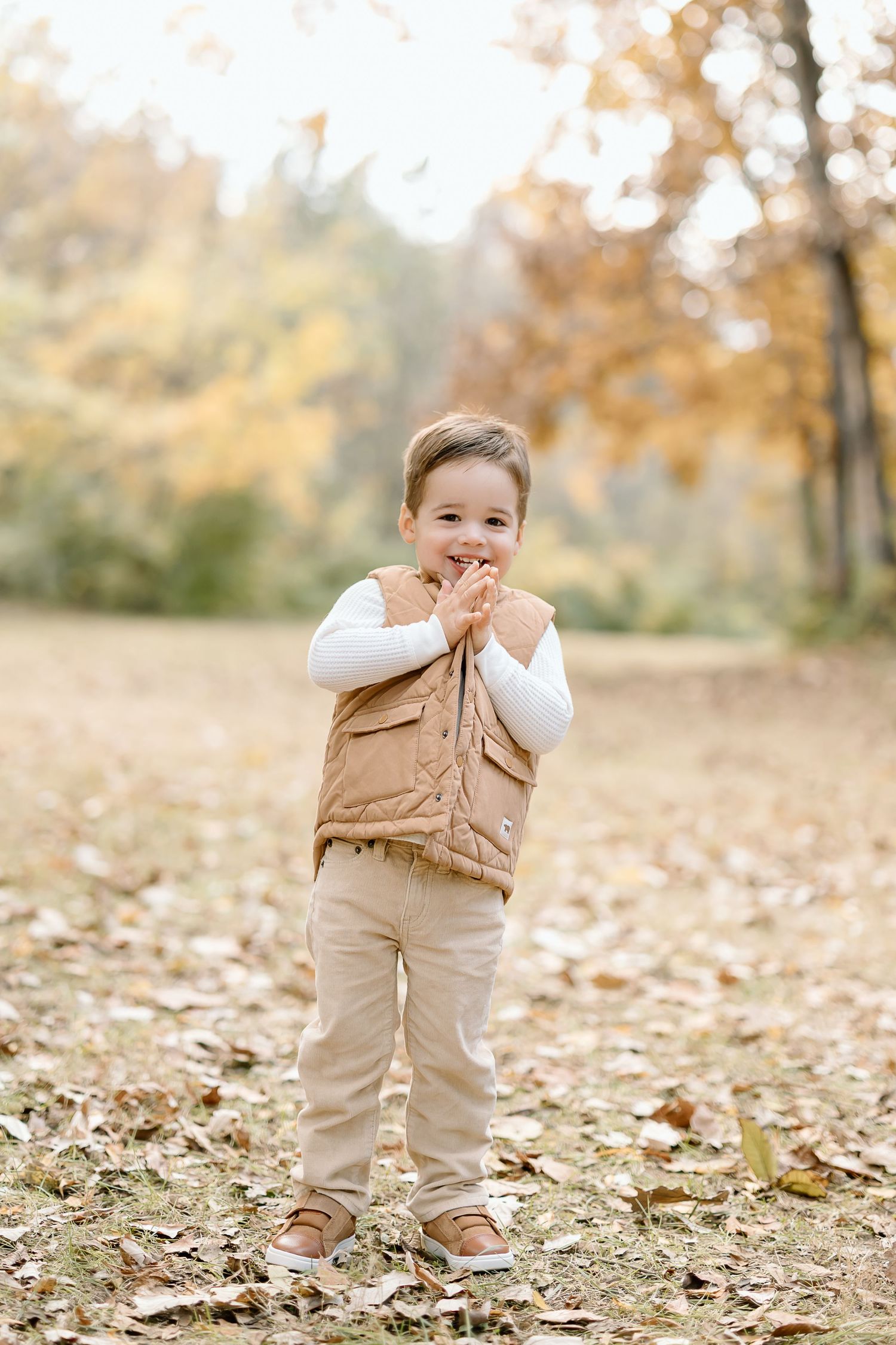 Young child in beige outfit plays joyfully among fallen autumn leaves on the ground during a fall photo session.