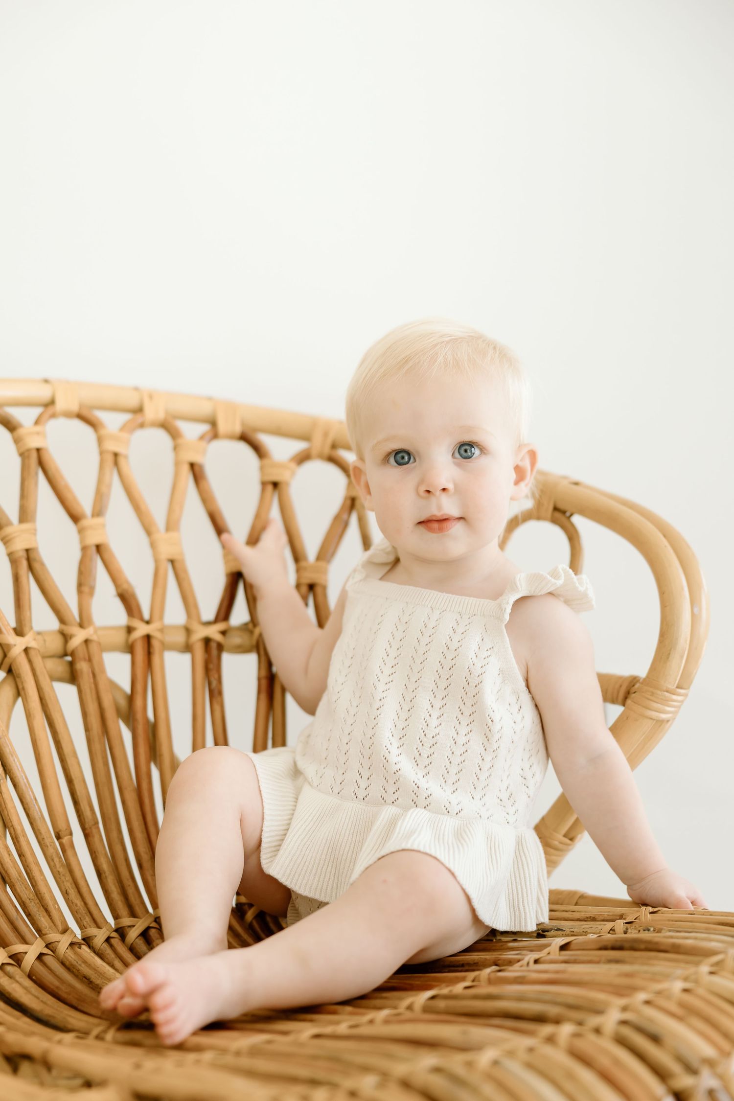 A baby in a white knit romper poses on a curved rattan chair in a series of lifestyle portraits.