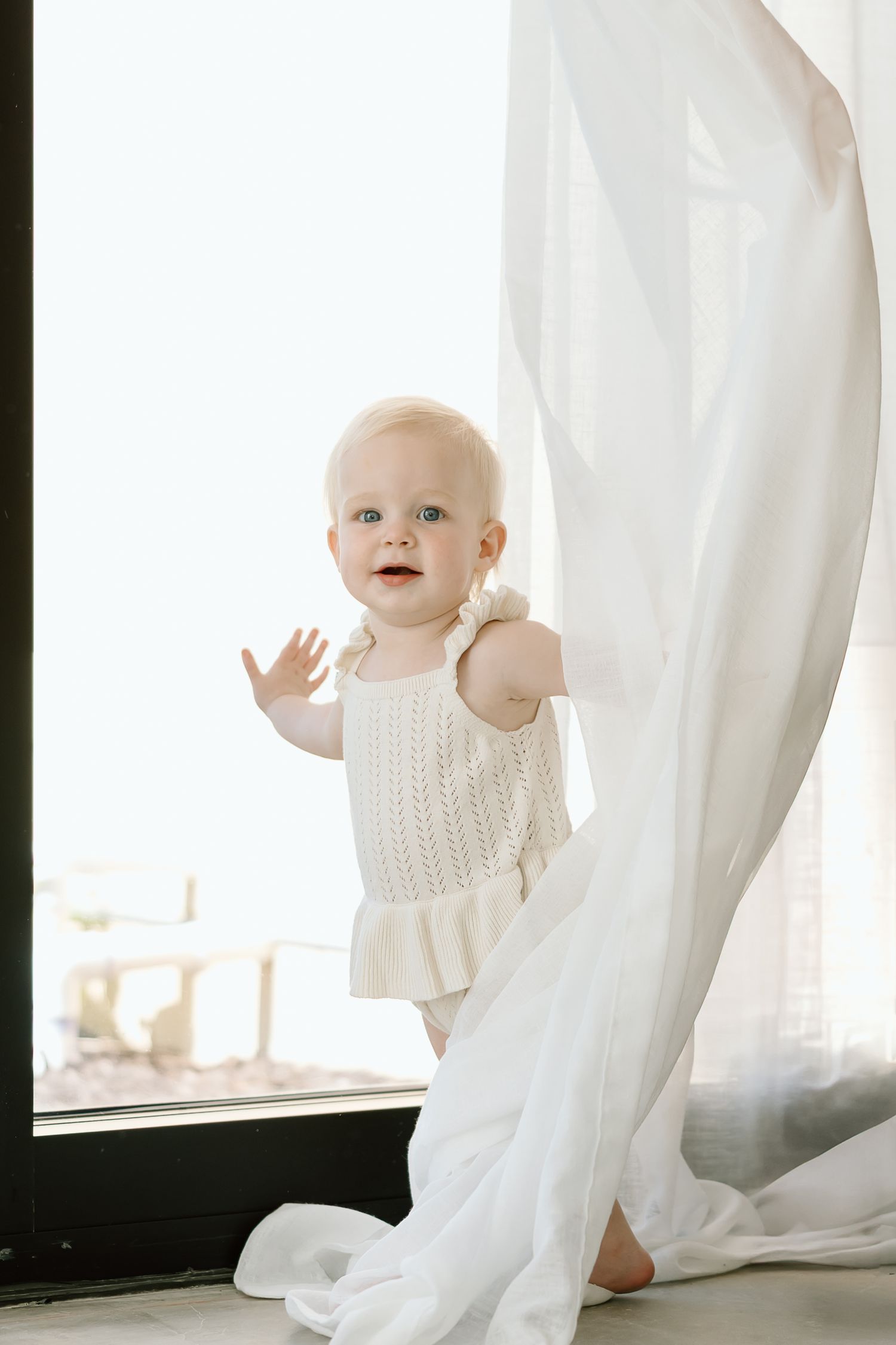 Baby in white knit sweater plays with sheer white curtains in bright sunlit room.