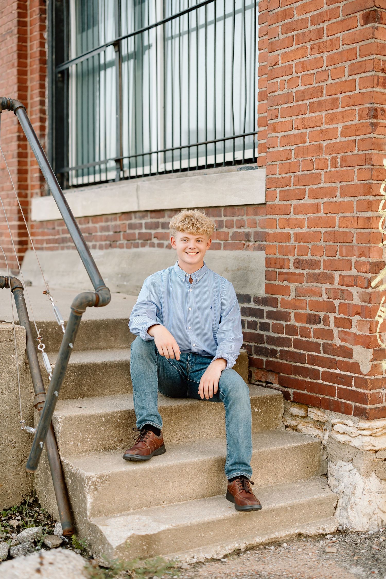 A person sits casually on concrete steps against a red brick wall wearing jeans and a light blue shirt.