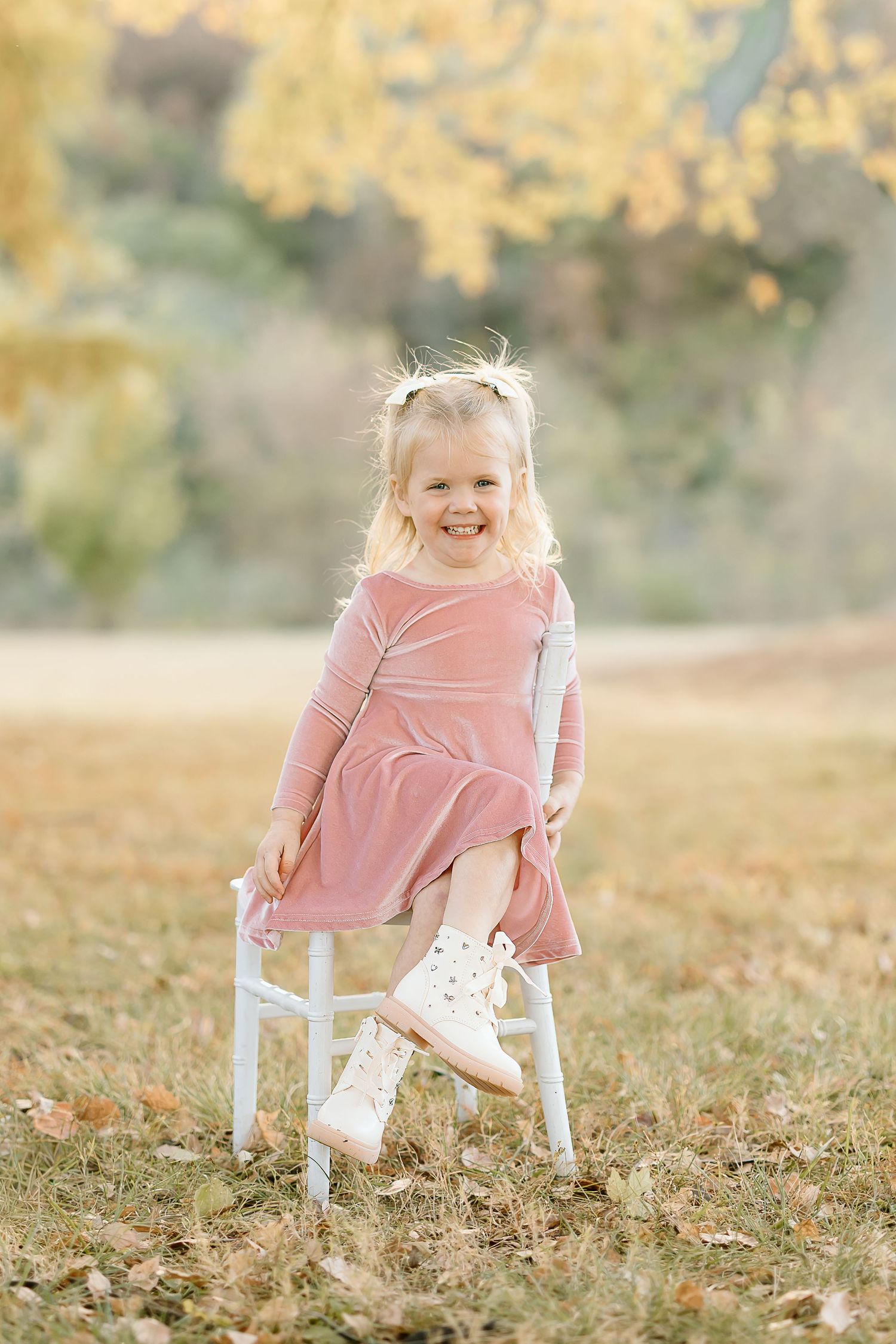 A small child in a pink dress sits in a white chair outdoors during golden hour with autumn leaves scattered on the ground.
