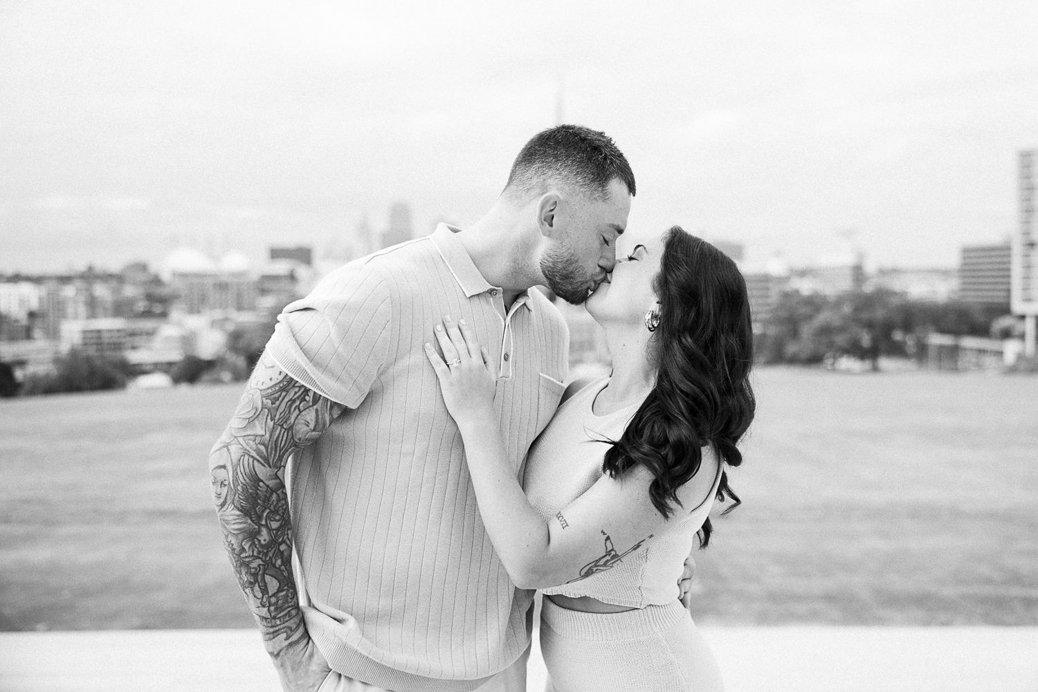 A couple embraces in a romantic outdoor photoshoot with a city skyline backdrop.