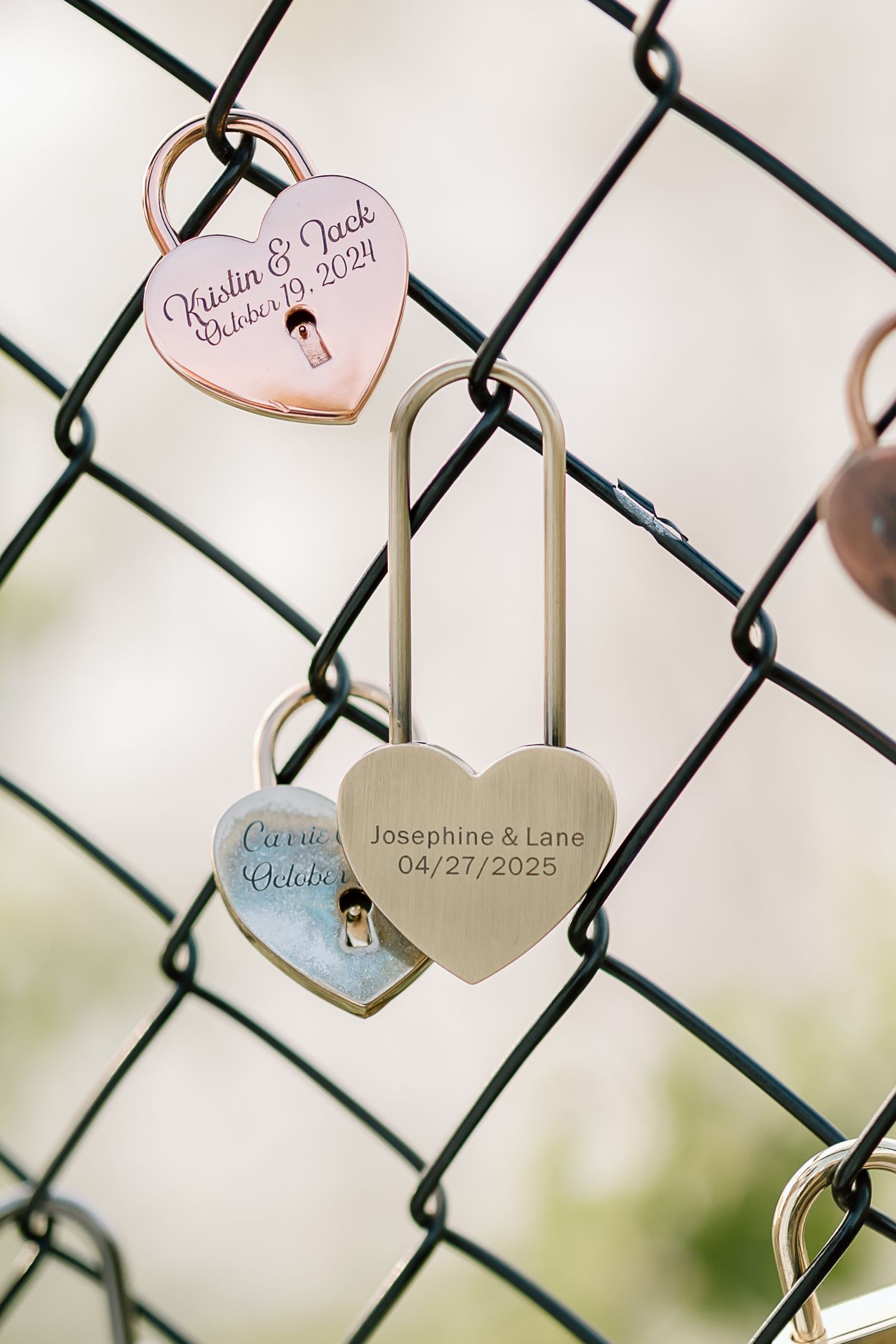 Love locks with engraved names and dates hang on a chain link fence in soft focus.