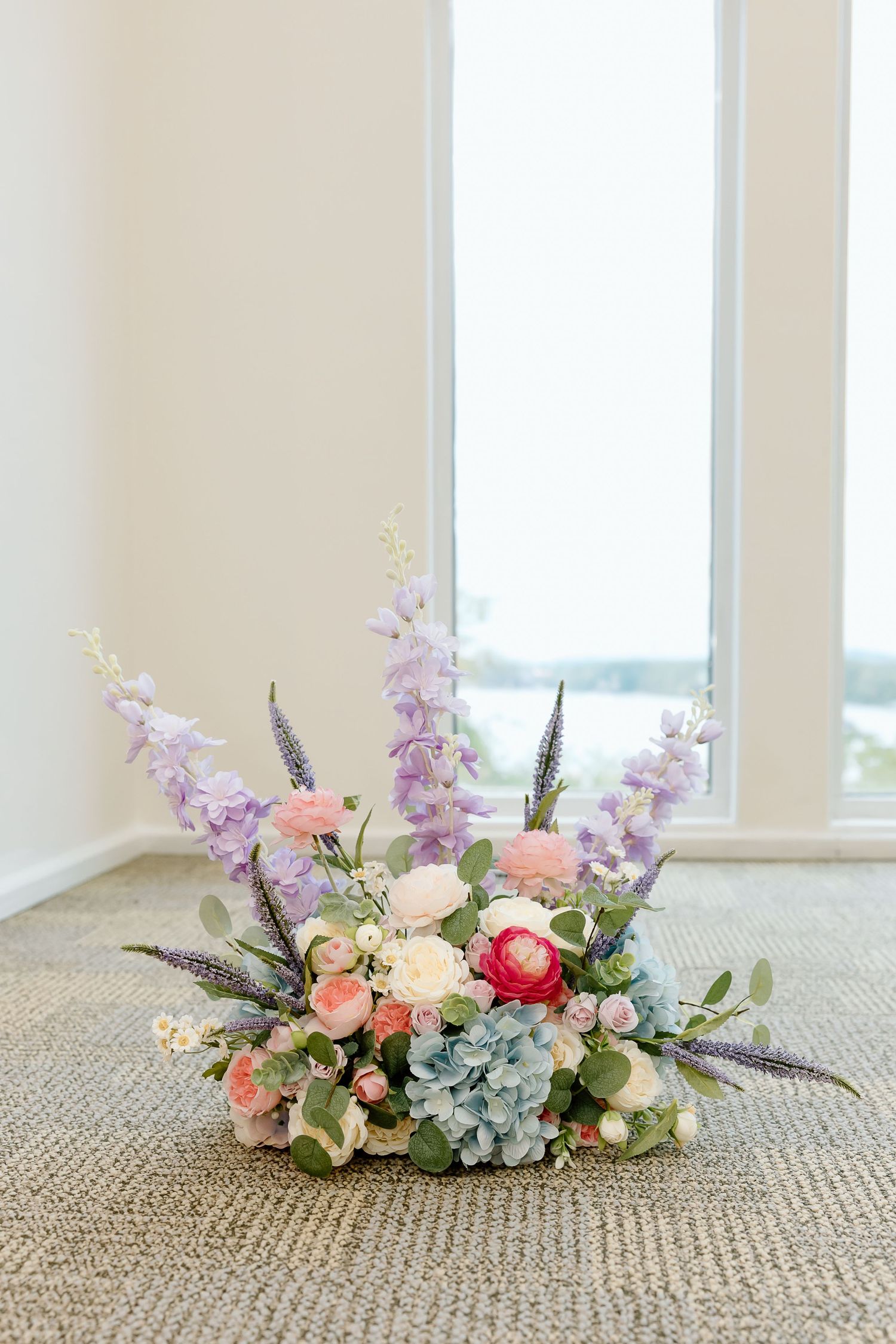 A delicate floral arrangement with pink and blue hydrangeas, roses, and greenery displayed on a beige carpet near windows.