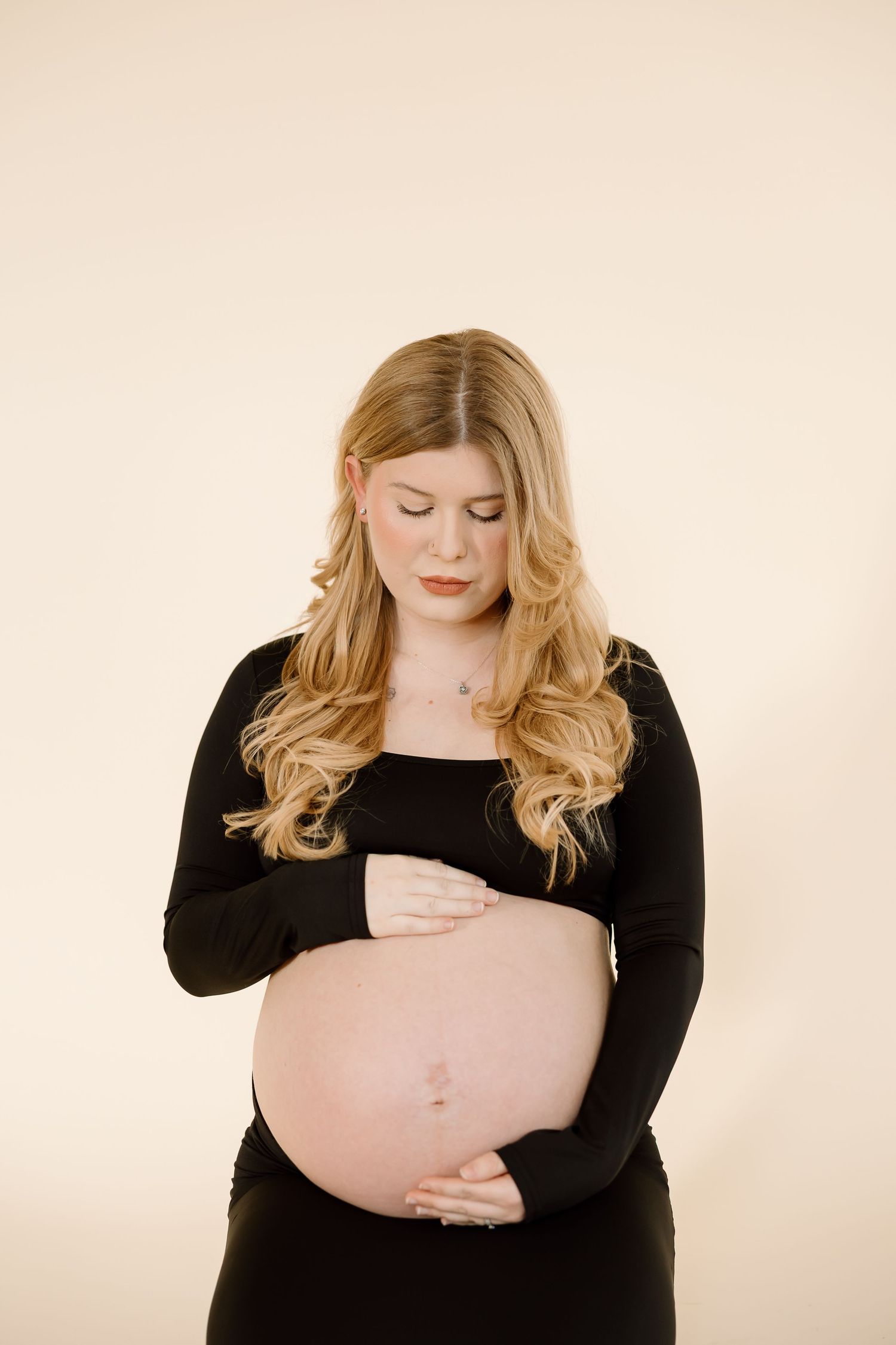 A pregnant woman in a black long sleeve dress poses against a beige background while holding her belly.