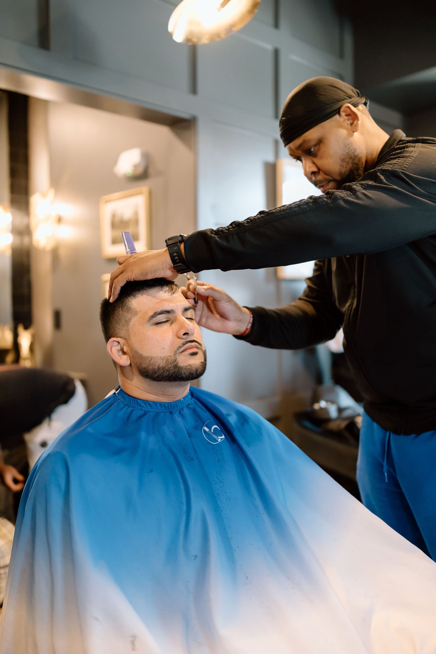 A barber in a black outfit carefully styles a client's hair in a modern barbershop, client wearing a blue cape.
