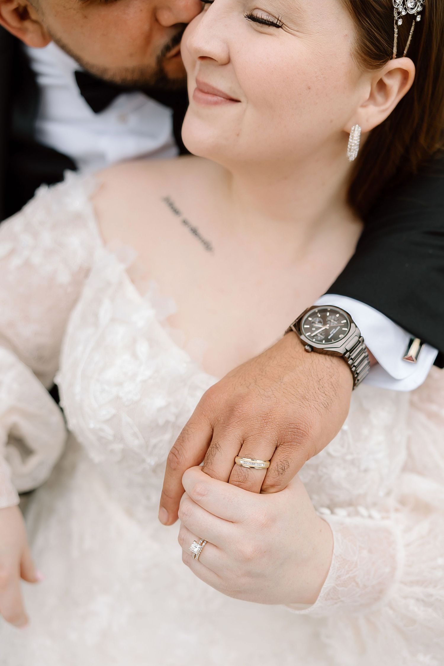 Bride and groom holding hands, showcasing wedding rings and watch, as groom kisses bride's cheek on wedding day.