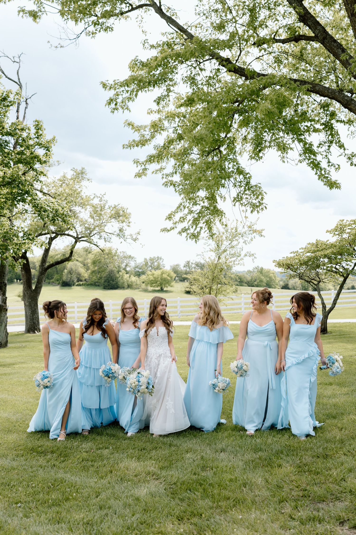 Bride in white gown walks with five bridesmaids in light blue dresses holding bouquets on a green lawn.