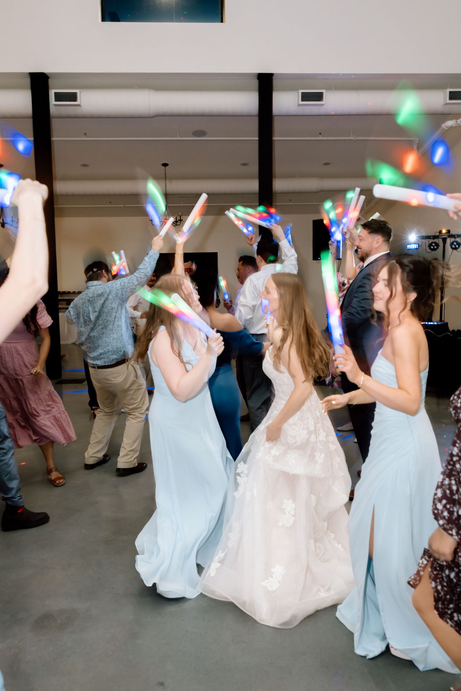 Bride in white gown dances with bridesmaids in light blue dresses, guests wave colorful LED wands at wedding reception.