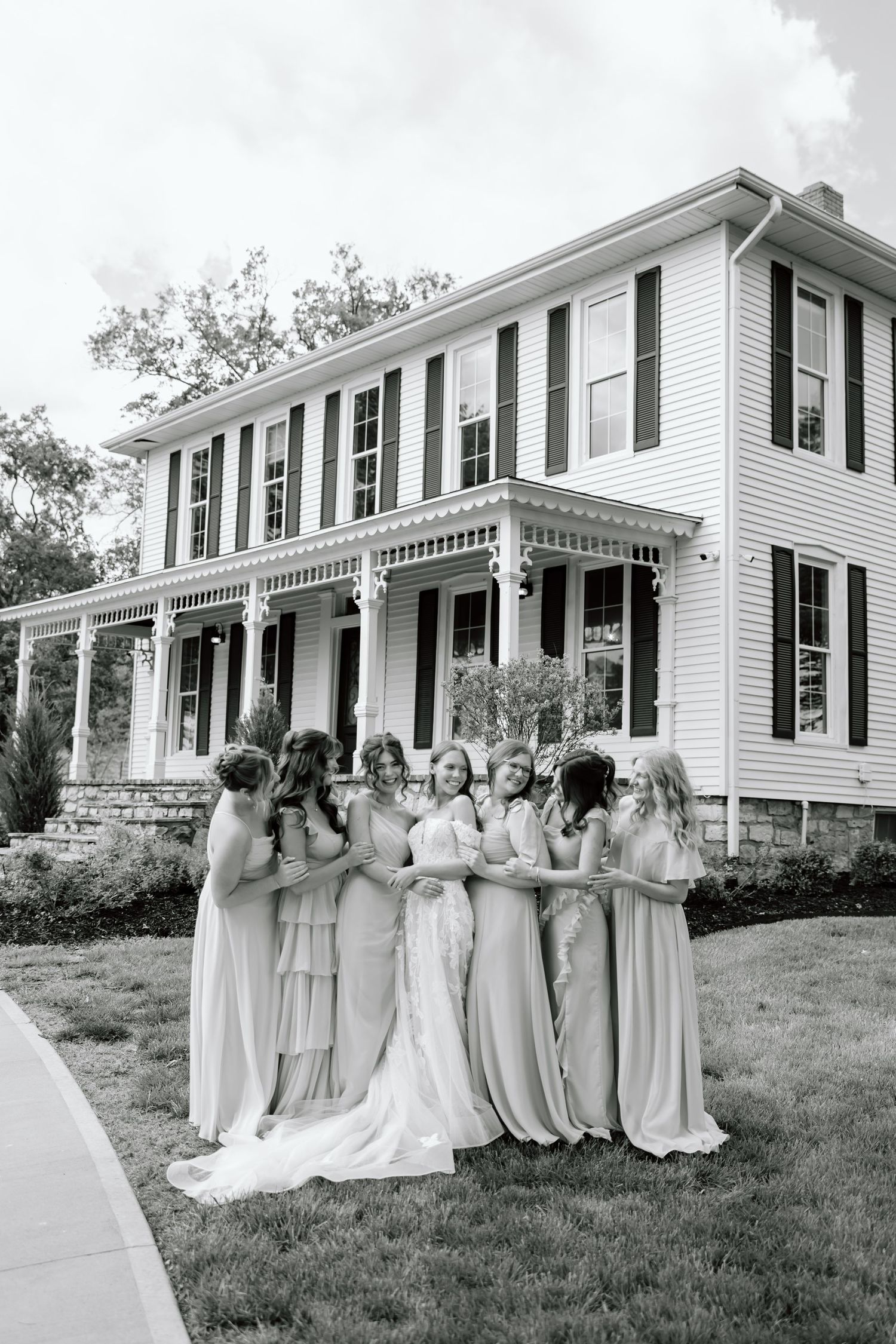 Bride and bridesmaids in elegant gowns posing together in front of a classic white colonial home.
