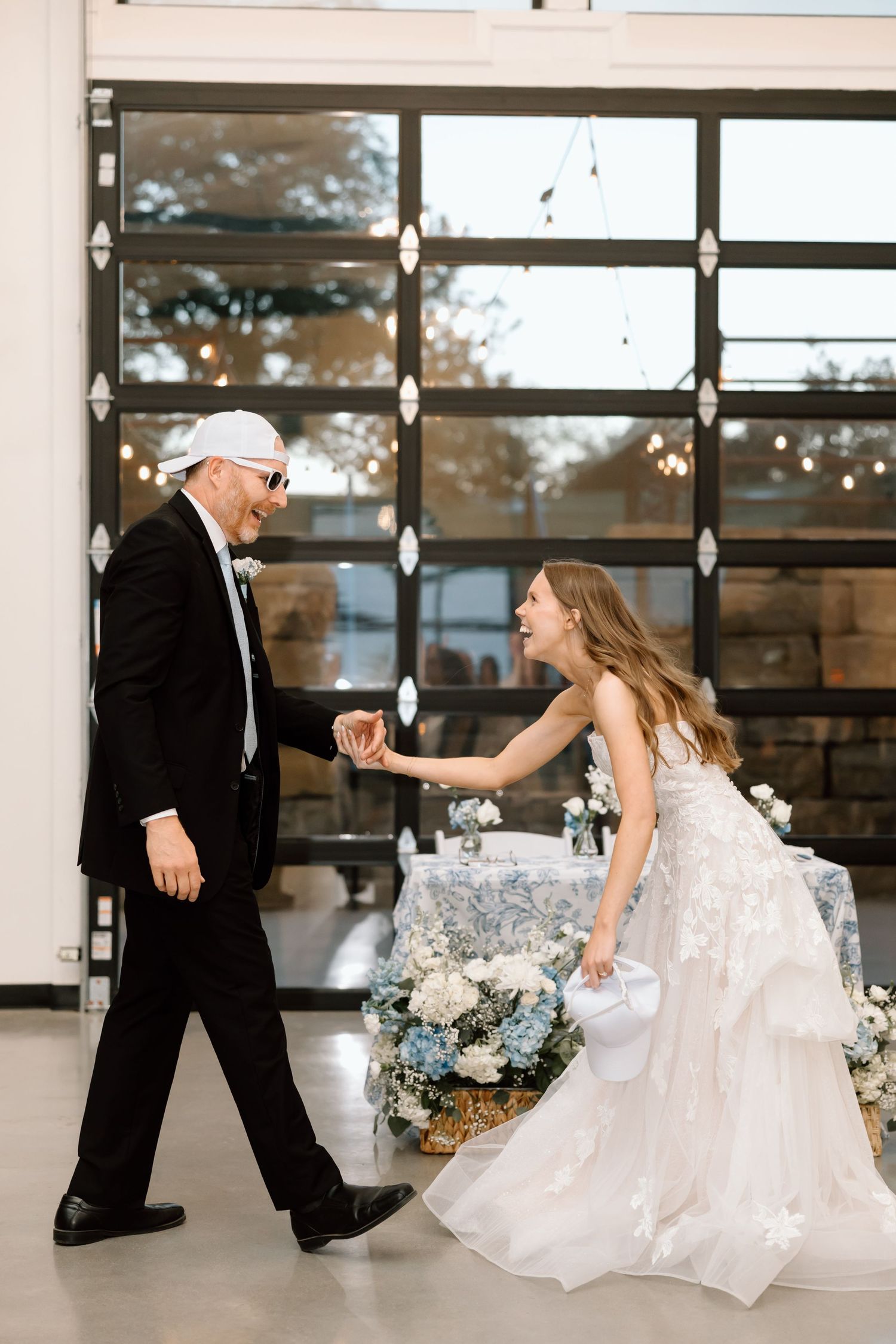 A flower girl reaches out to an older man in a tuxedo and sunglasses dancing near floral arrangements at a wedding.