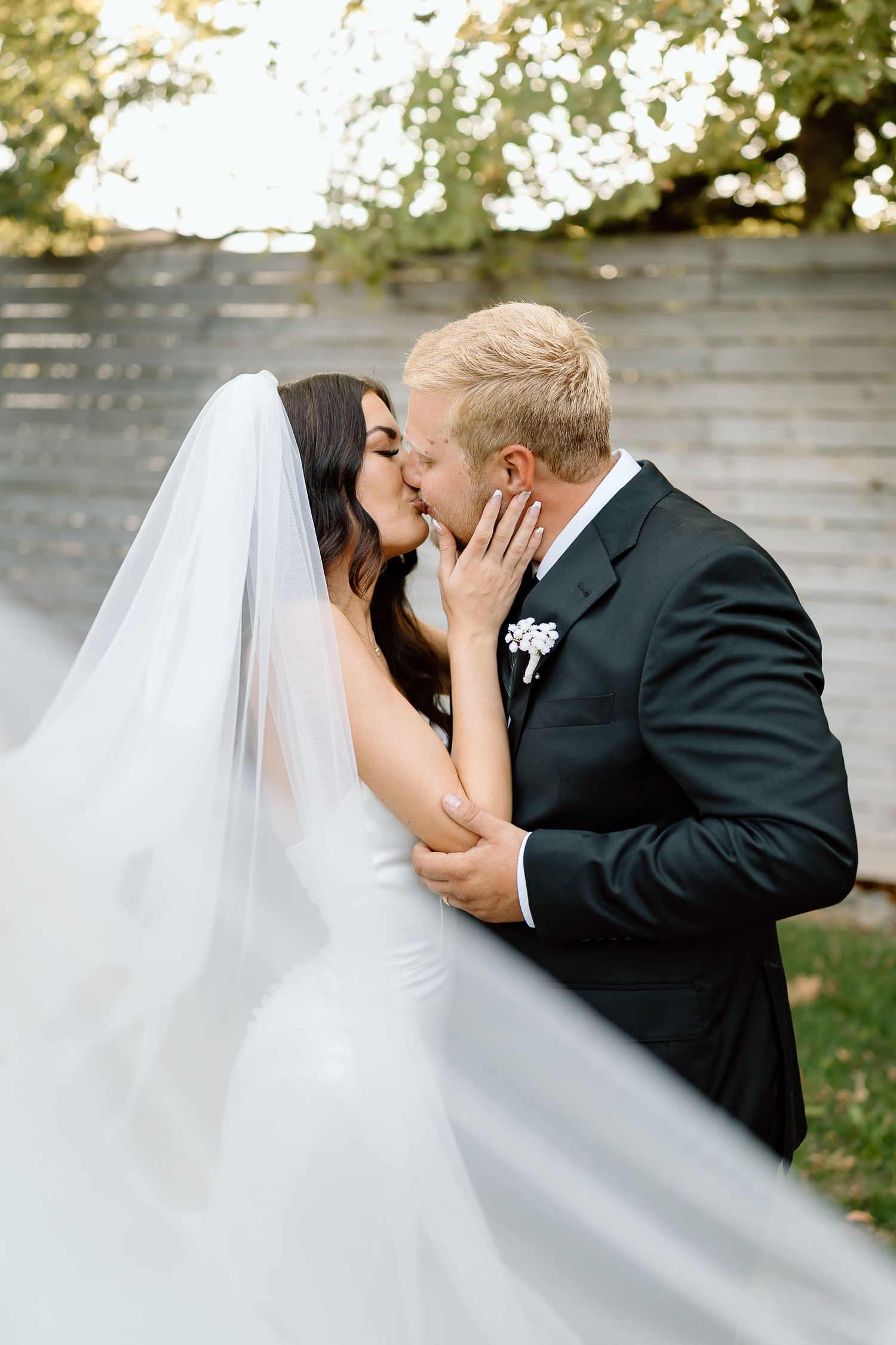 A romantic wedding kiss moment between a bride in white and groom in black suit against a soft bokeh background.