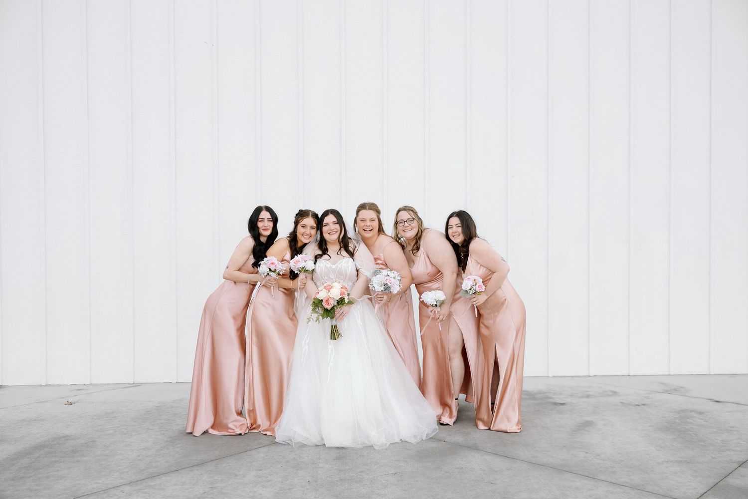 Wedding party wearing matching blush pink bridesmaid dresses poses together against white wall.